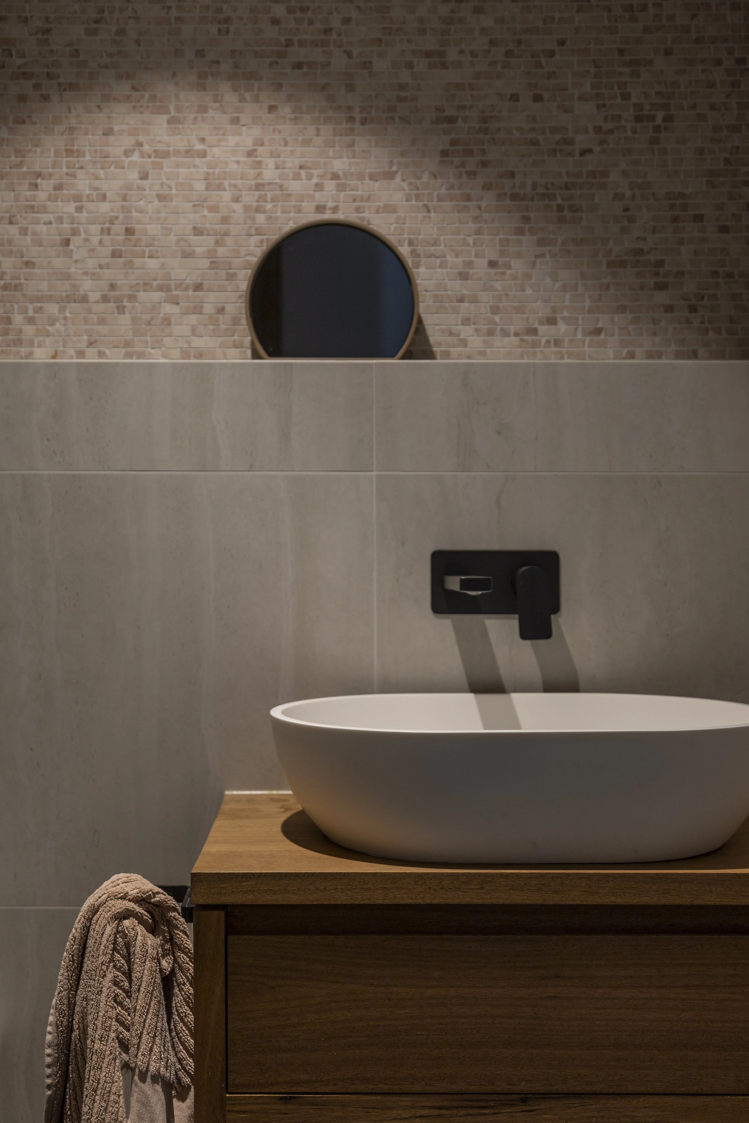 Modern bathroom with a white vessel sink on a wooden vanity, beige tiled wall, a round mirror, and a beige towel hanging on the side.