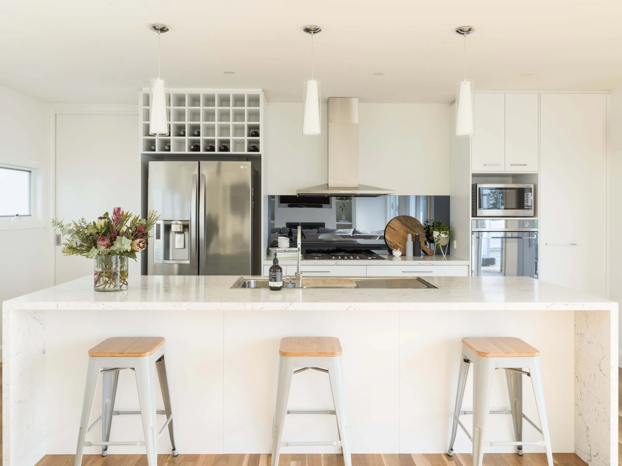 A modern kitchen with a white marble island, four light wood barstools, a vase of flowers, and a view into the living room with a TV and sofa.