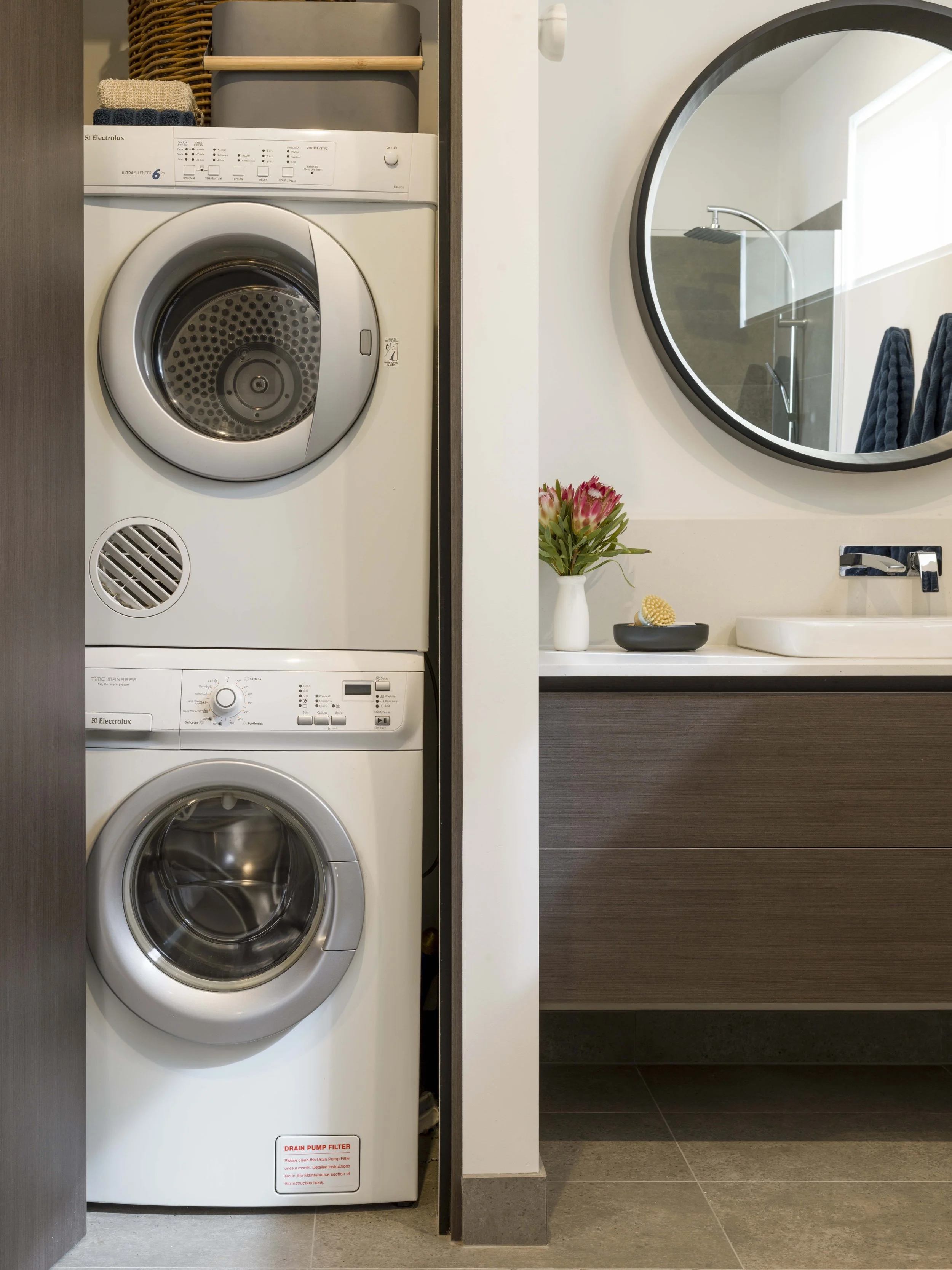 A washer and dryer stacked in a laundry area, with a decorative flower arrangement and bathroom mirror on the right.