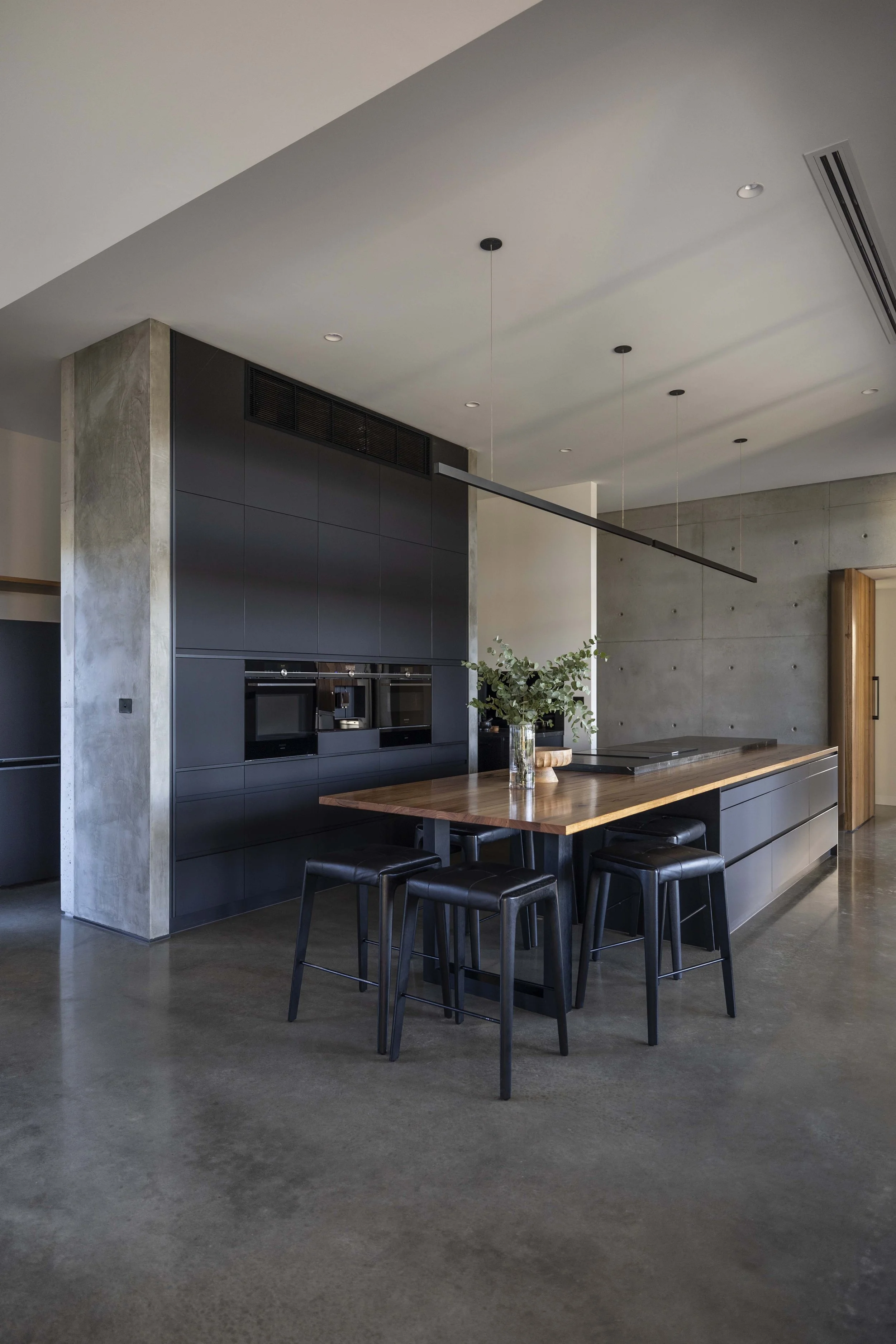 Modern kitchen with black cabinetry, a wooden island, and barstools, featuring concrete columns and gray walls.