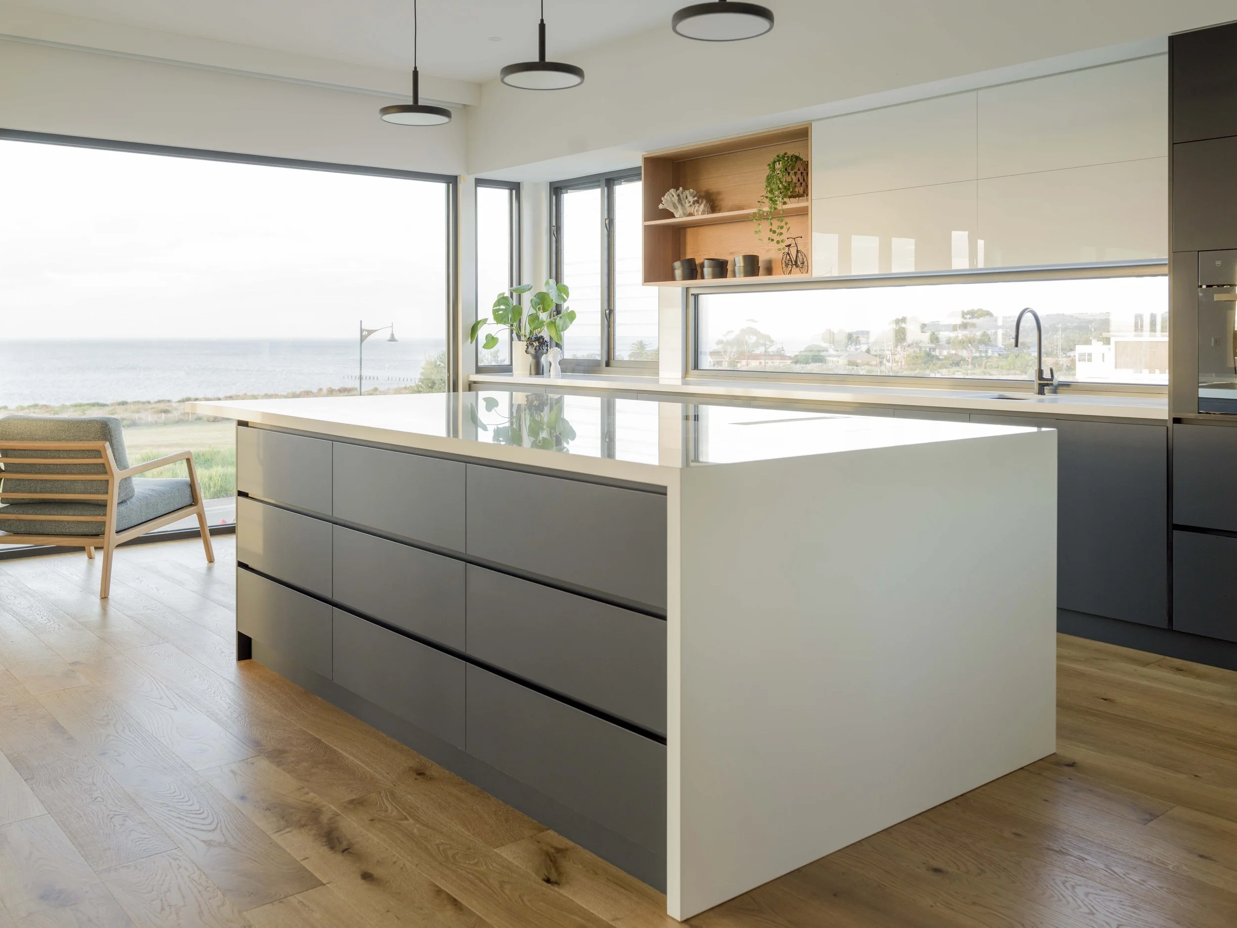 Modern kitchen with large window overlooking the ocean, featuring a white island, plant, and minimal decor.