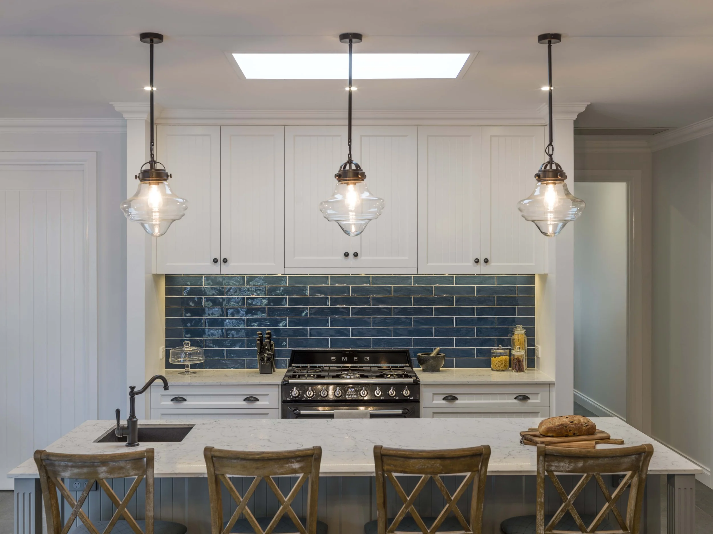 Modern kitchen with white cabinets, blue tiled backsplash, three pendant lights, a black stove, and a marble island with a loaf of bread and wooden chairs.