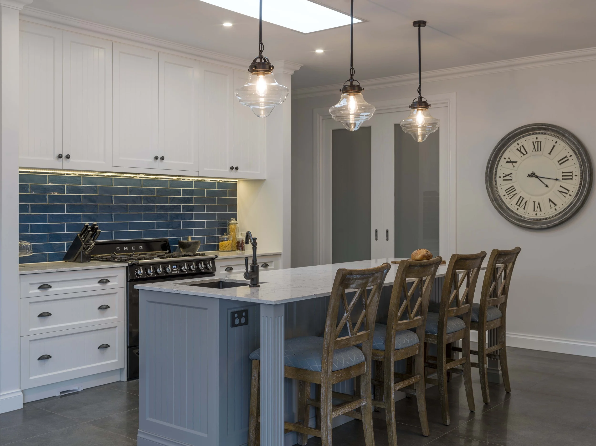 A modern kitchen with white cabinets, a blue tile backsplash, a stainless steel stove, and a white marble island with a built-in power outlet. There are wooden chairs with patterned cushions around the island and three pendant lights hanging from the