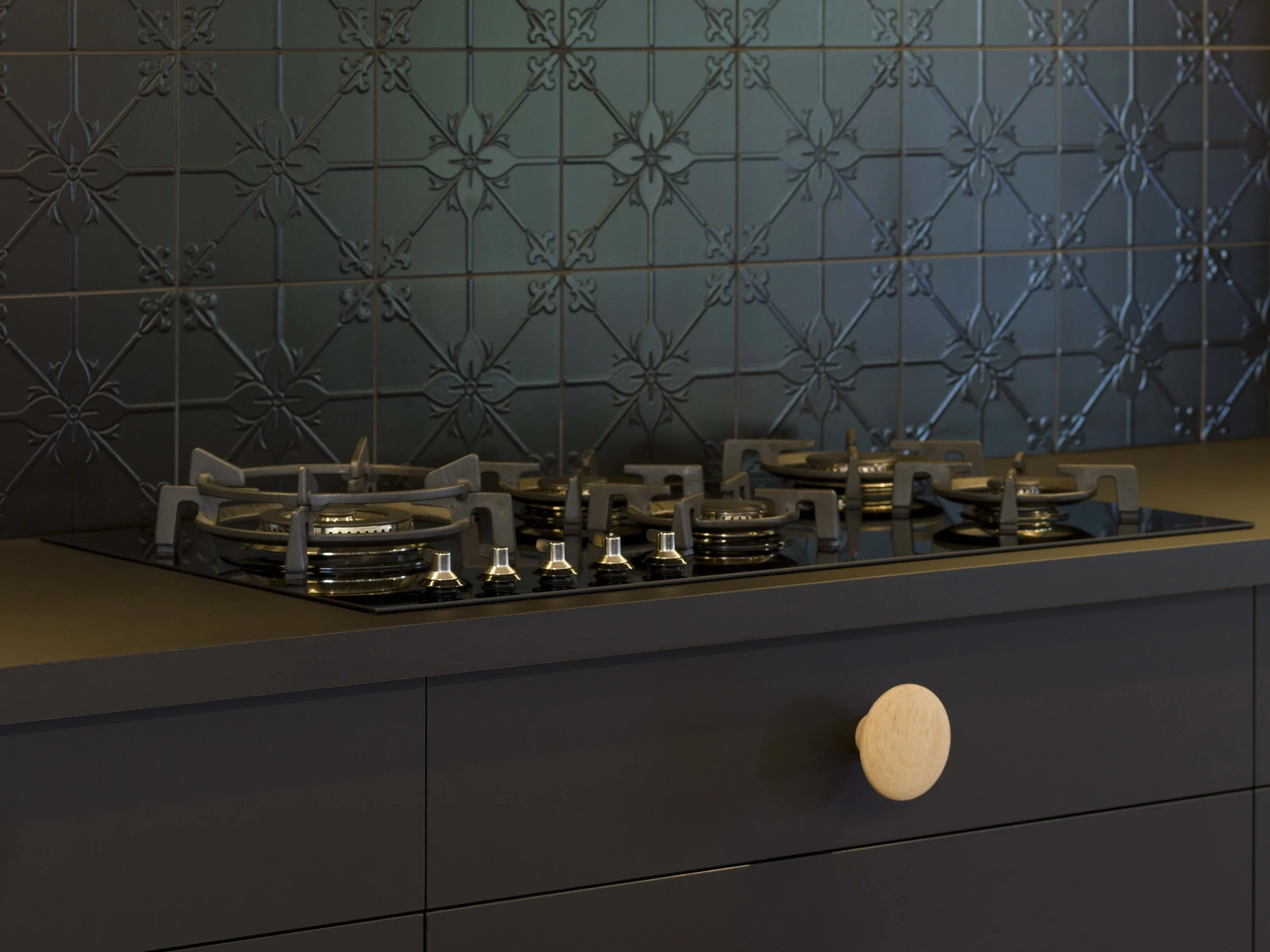 Black kitchen backsplash with decorative floral tile pattern, gas stove with four burners, and a wooden spoon resting on the counter.