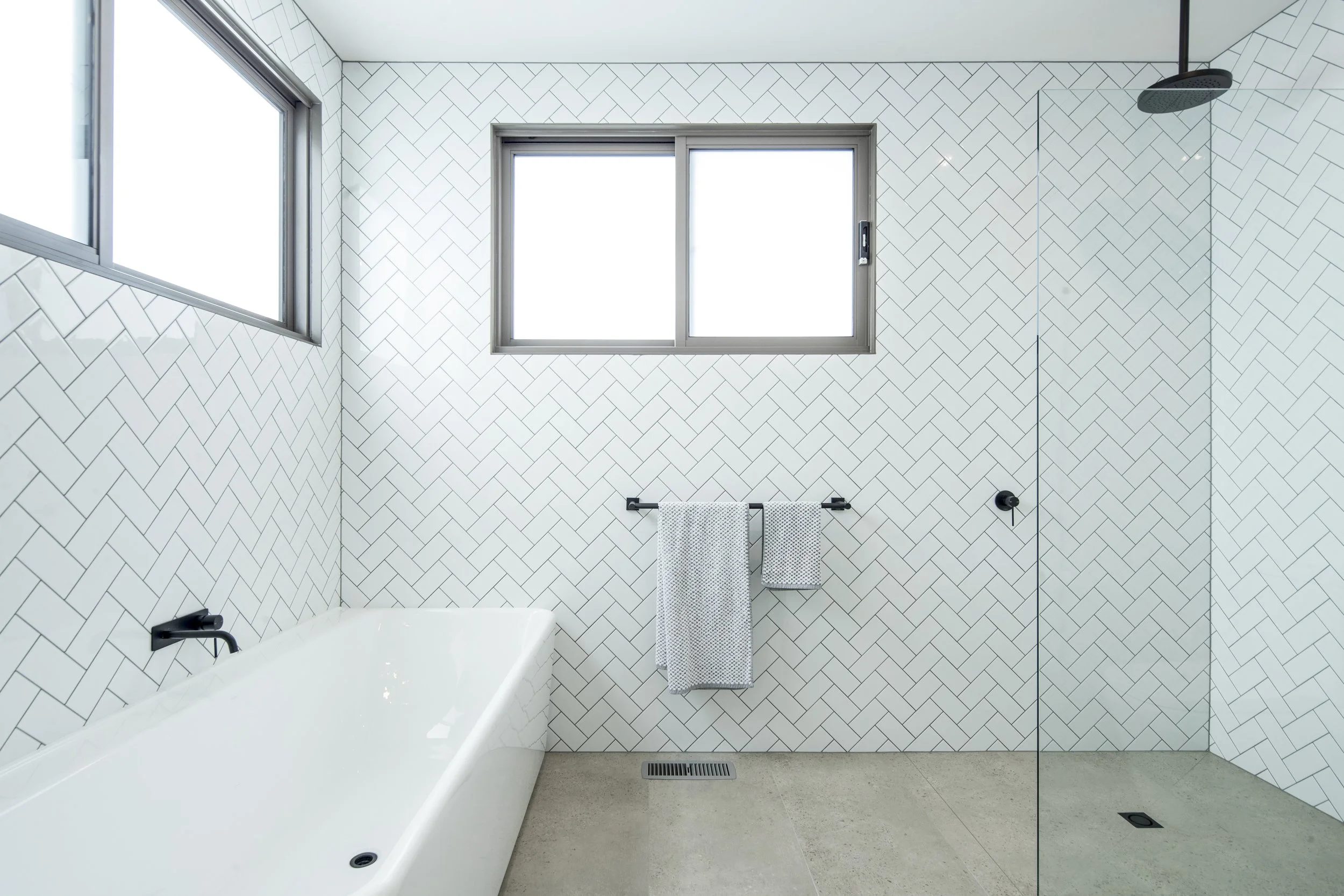 Bathroom with white herringbone tiled walls, window, bathtub with black fixtures, and a walk-in shower with a glass door.