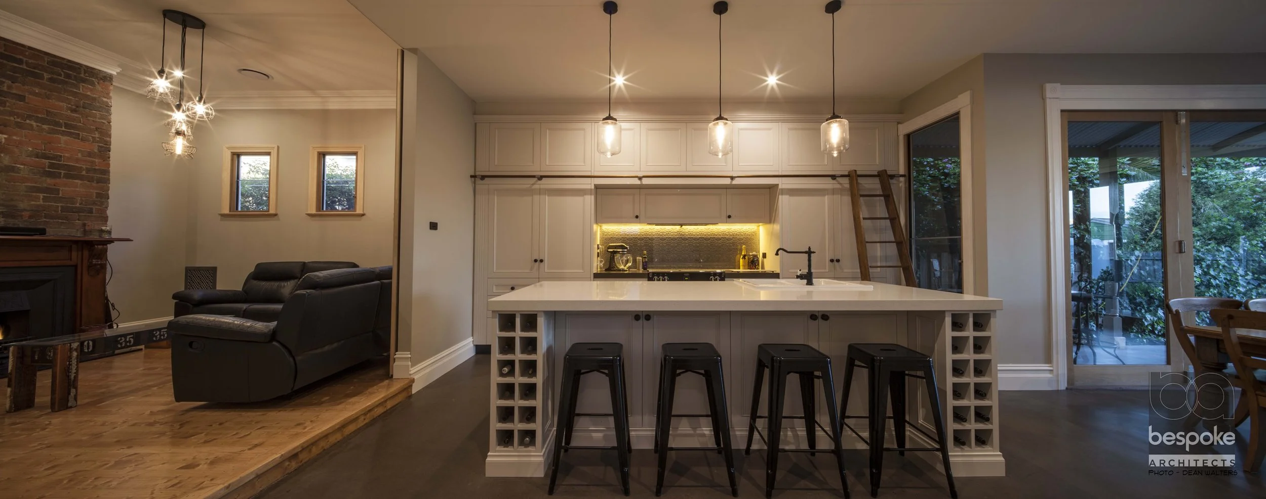 Modern kitchen with white cabinetry, island with wine rack sides, black barstools, pendant lighting, adjacent dining area, and sliding glass door leading to outdoor patio.