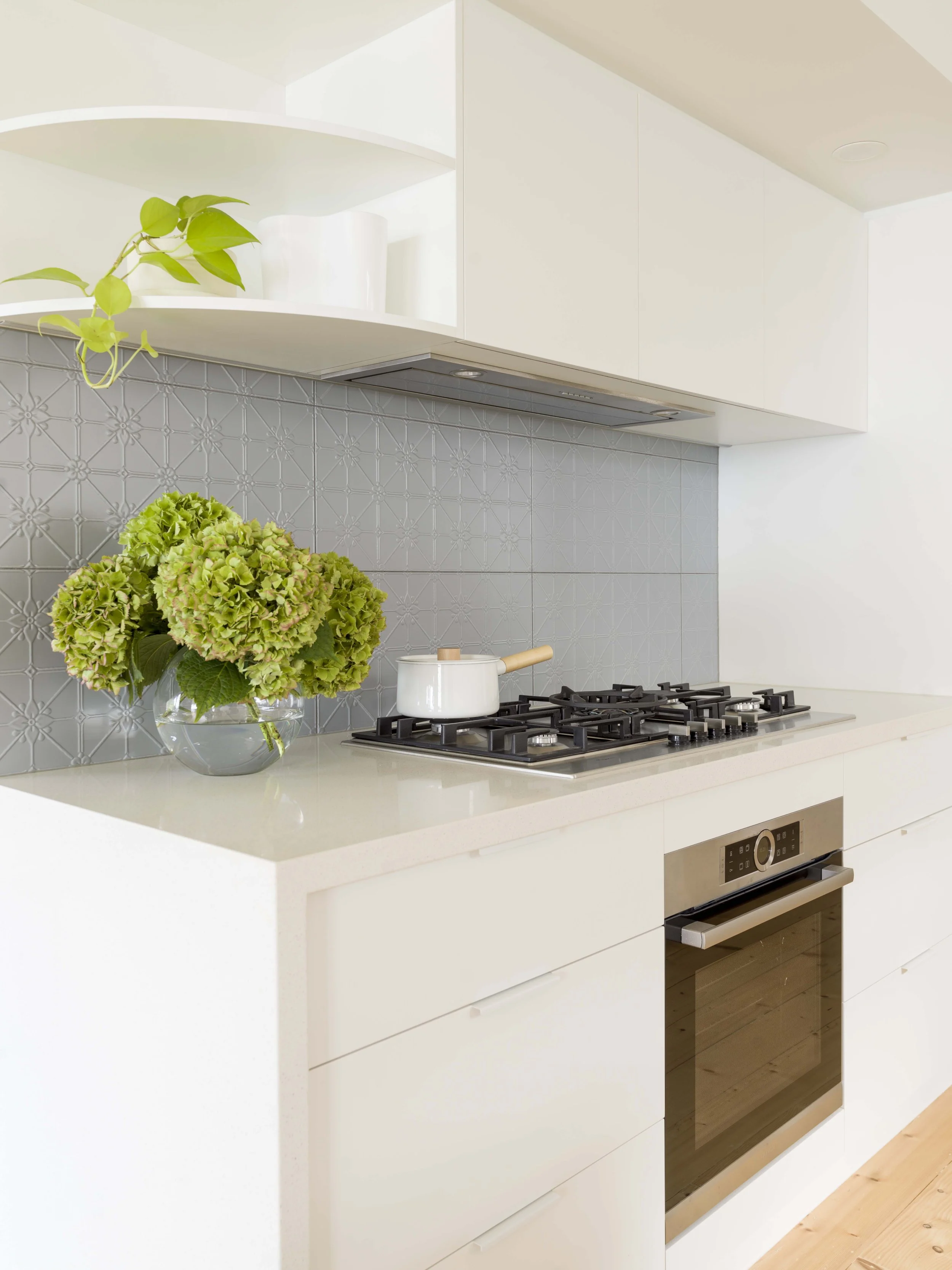 Modern kitchen with white cabinetry, a gas stove with a white pot, a vase with green hydrangeas, and a patterned gray tile backsplash.