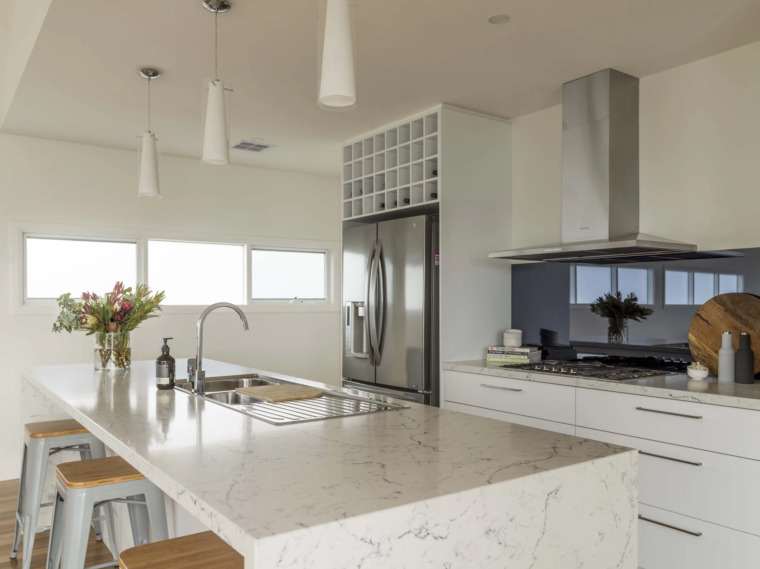 Modern kitchen with a white marble island, flowers, and high-end appliances including a stainless steel refrigerator and range hood, with two wooden stools at the island.