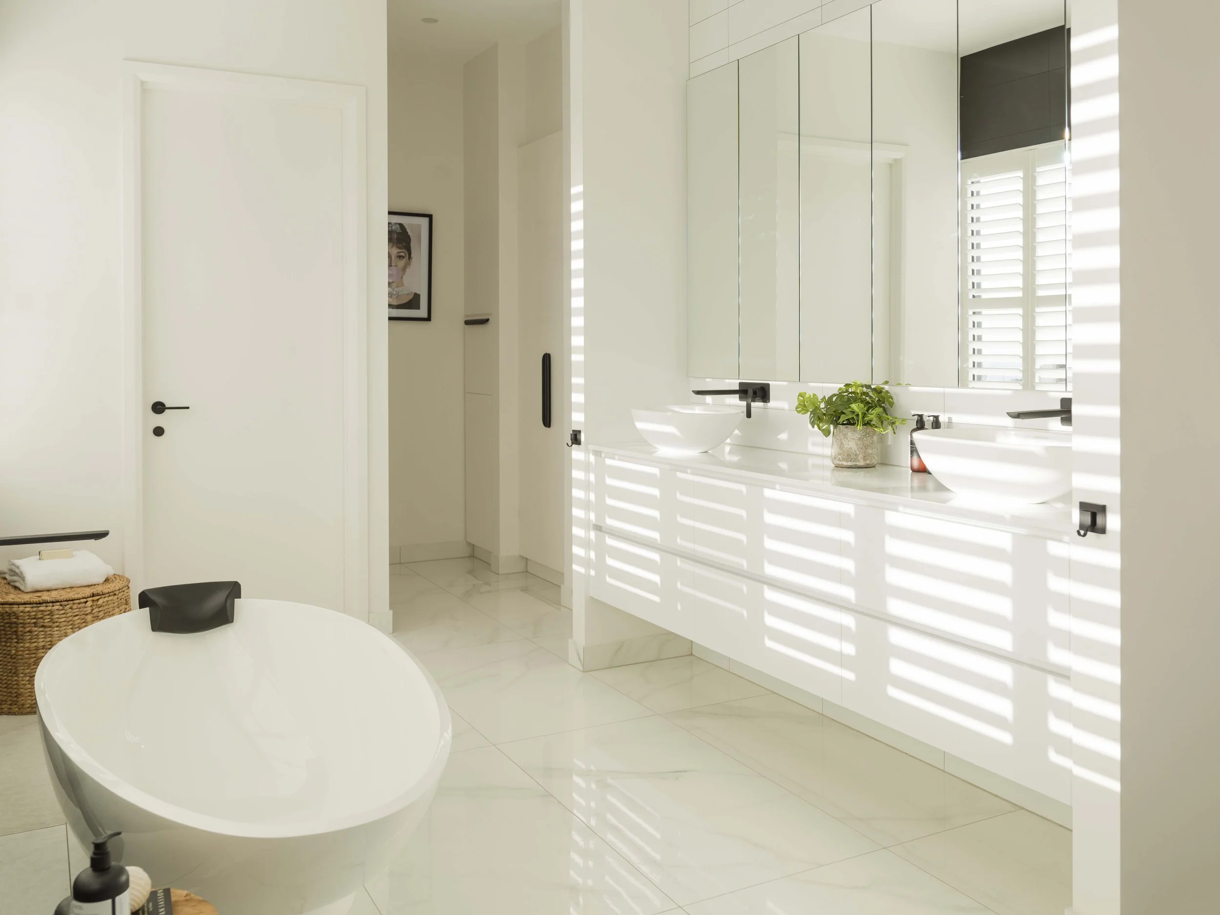 A modern bathroom with white walls and cabinets, a white countertop with a vessel sink, a large mirror, a plant, and sunlight through window shutters.