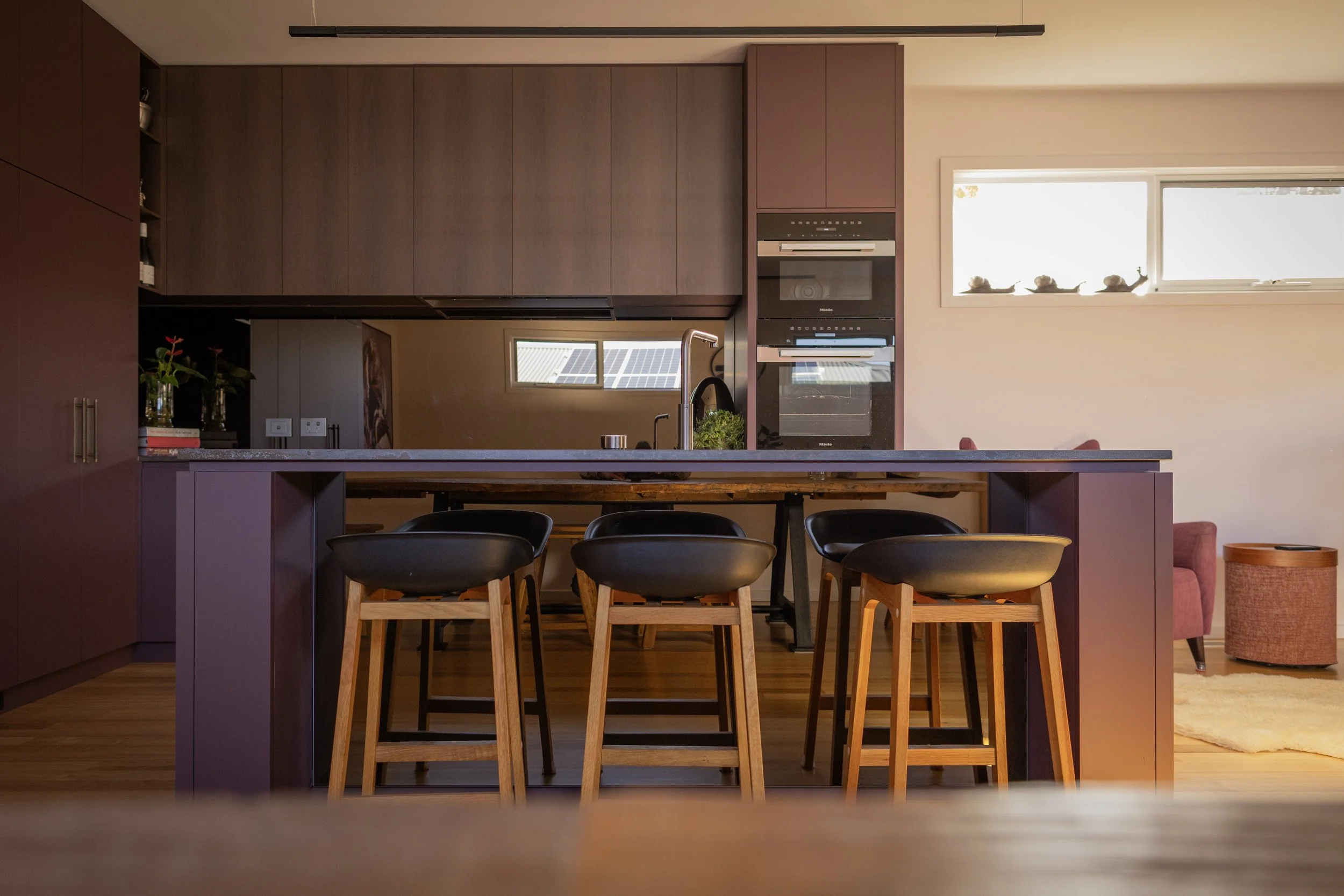 Interior view of a modern kitchen with purple cabinetry, wooden chairs around a kitchen island, black chairs, a window, and a small living area with a pink armchair and a round orange side table.