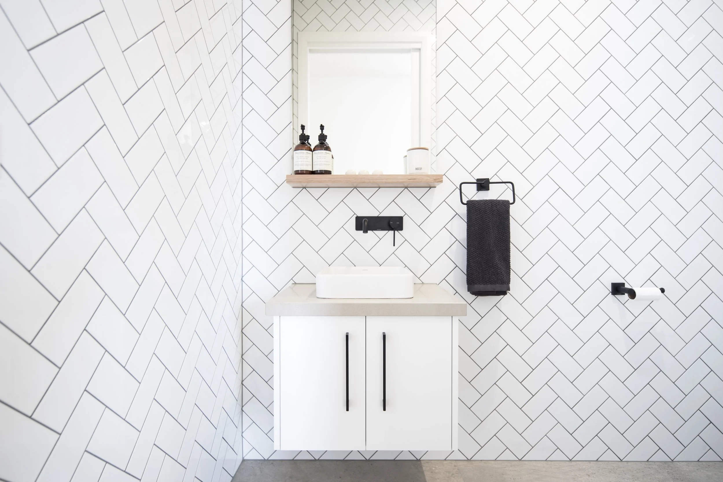 Modern bathroom with white herringbone tile walls, a white vanity with black handles, a rectangular vessel sink, a round mirror, and black fixtures including a towel holder and toilet paper holder.