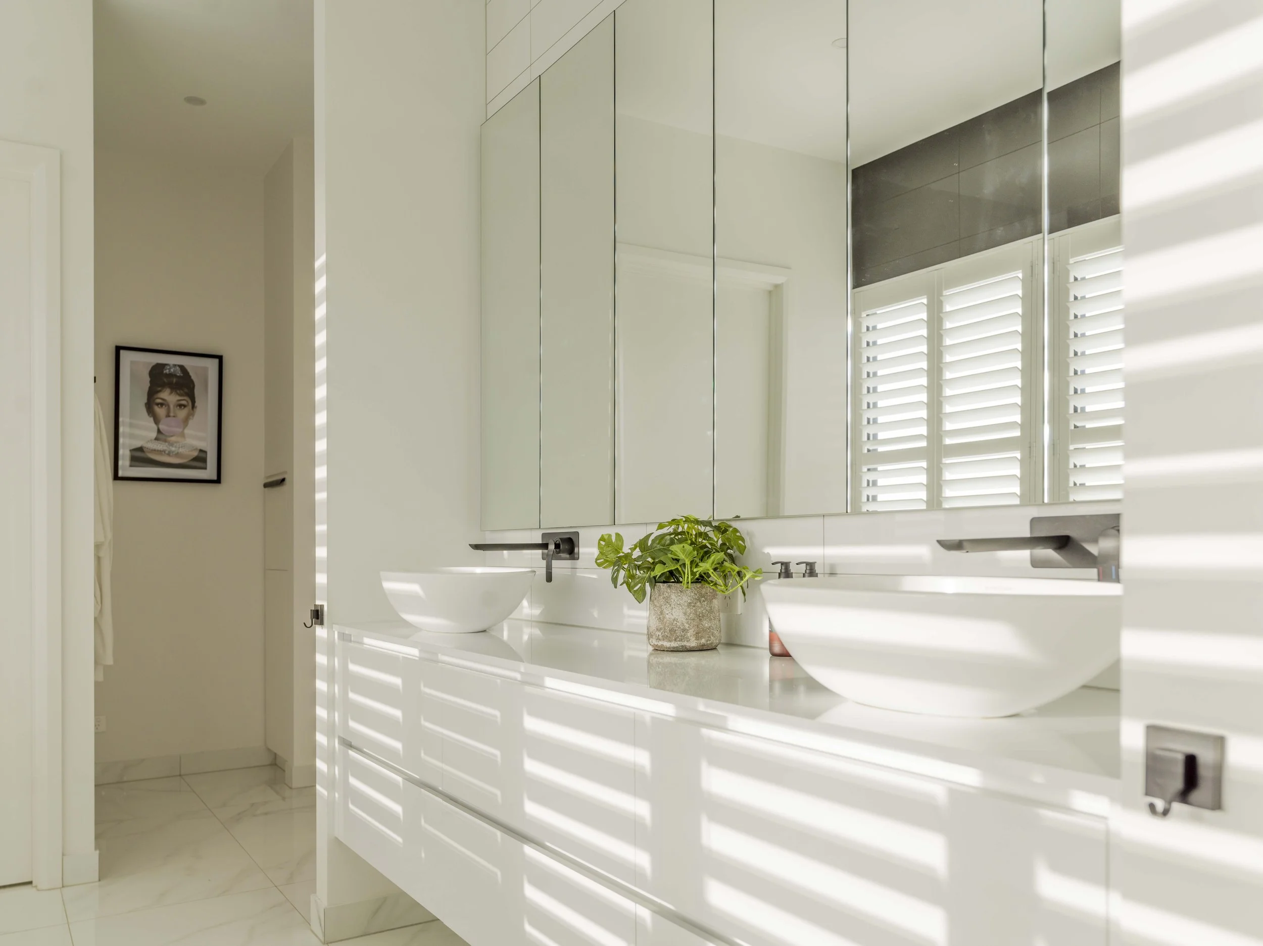 A modern bathroom with white cabinetry, a large mirror, a white vessel sink with a black faucet, a potted green plant, and plantation shutters on the windows, with sunlight casting shadows.