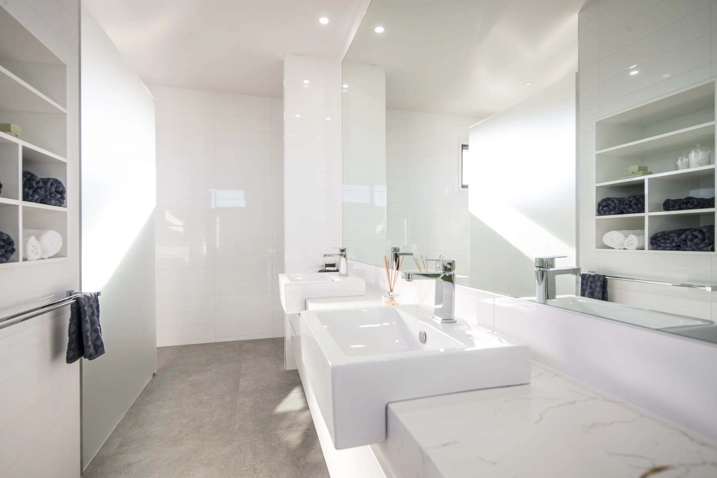 A modern white bathroom with two sinks, a large mirror, and open shelving with neatly rolled dark and white towels. The room is brightly lit with recessed ceiling lights.