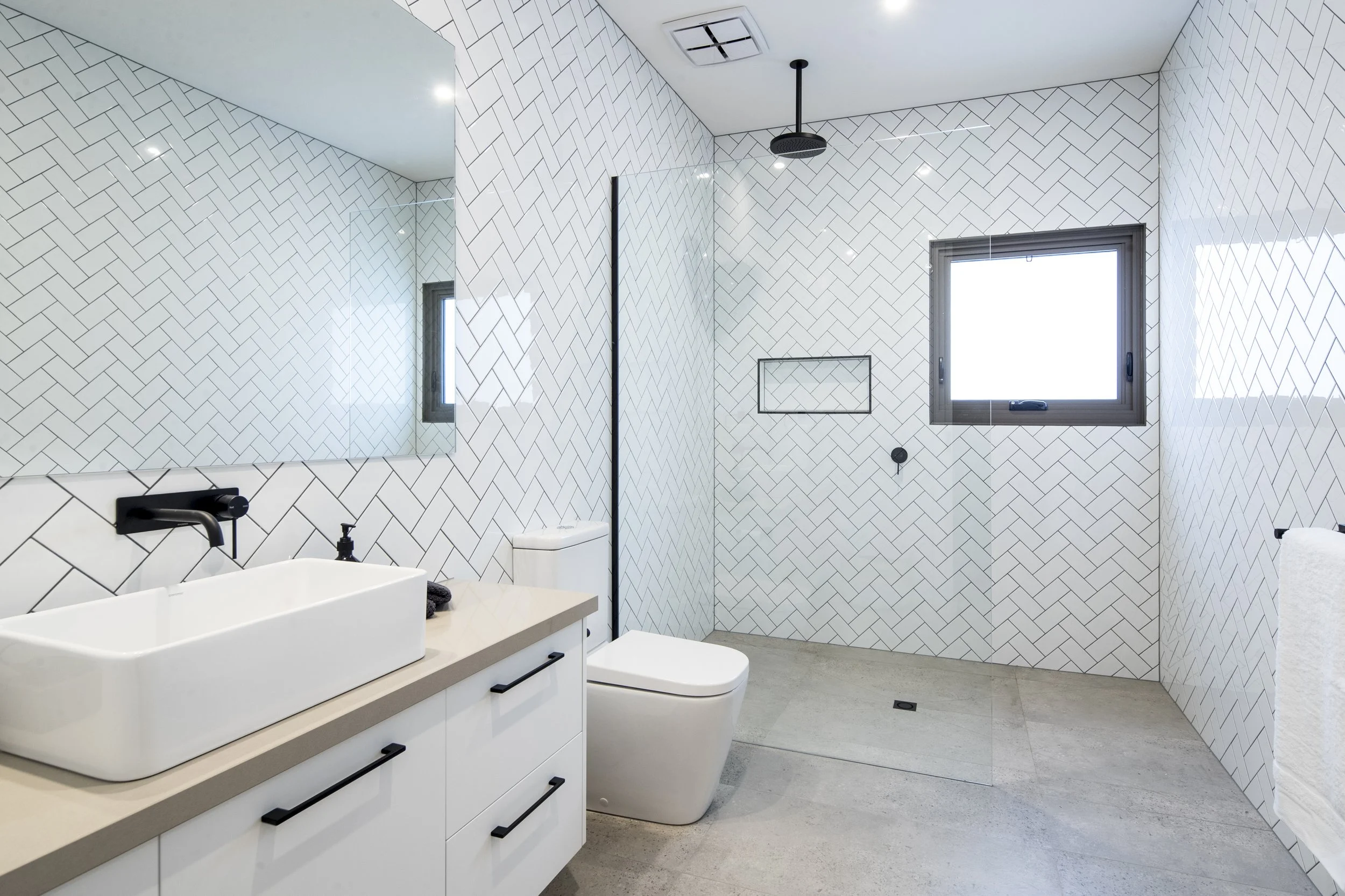 Modern bathroom with white herringbone tile walls, a large mirror, black fixtures, and a window with natural light.