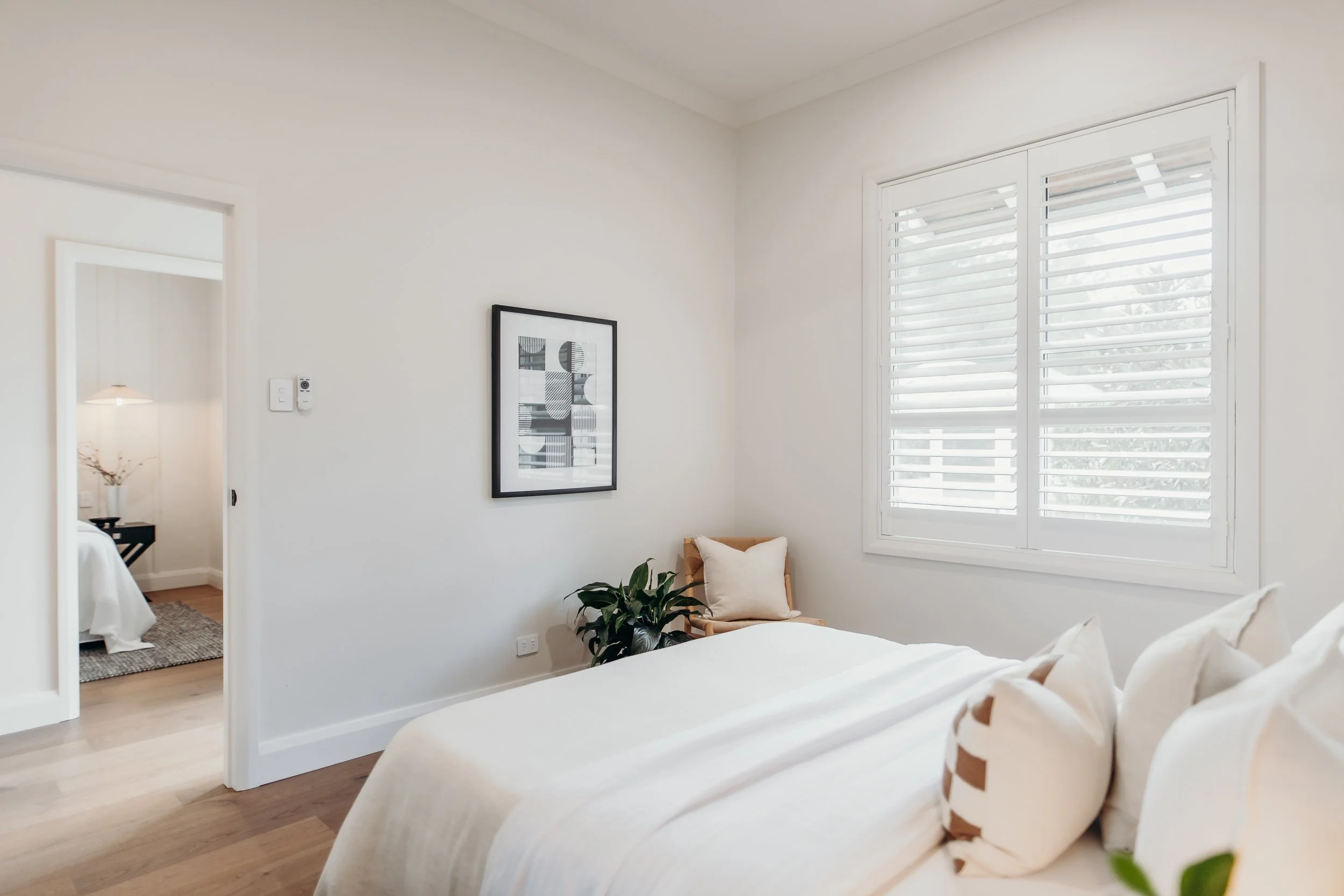 A bright bedroom with a bed, white bedding, and pillows. There is a window with white shutters, a framed abstract art piece on the wall, and a chair with a pillow and a houseplant. An adjoining room is visible with a bed, side table, and lamp.