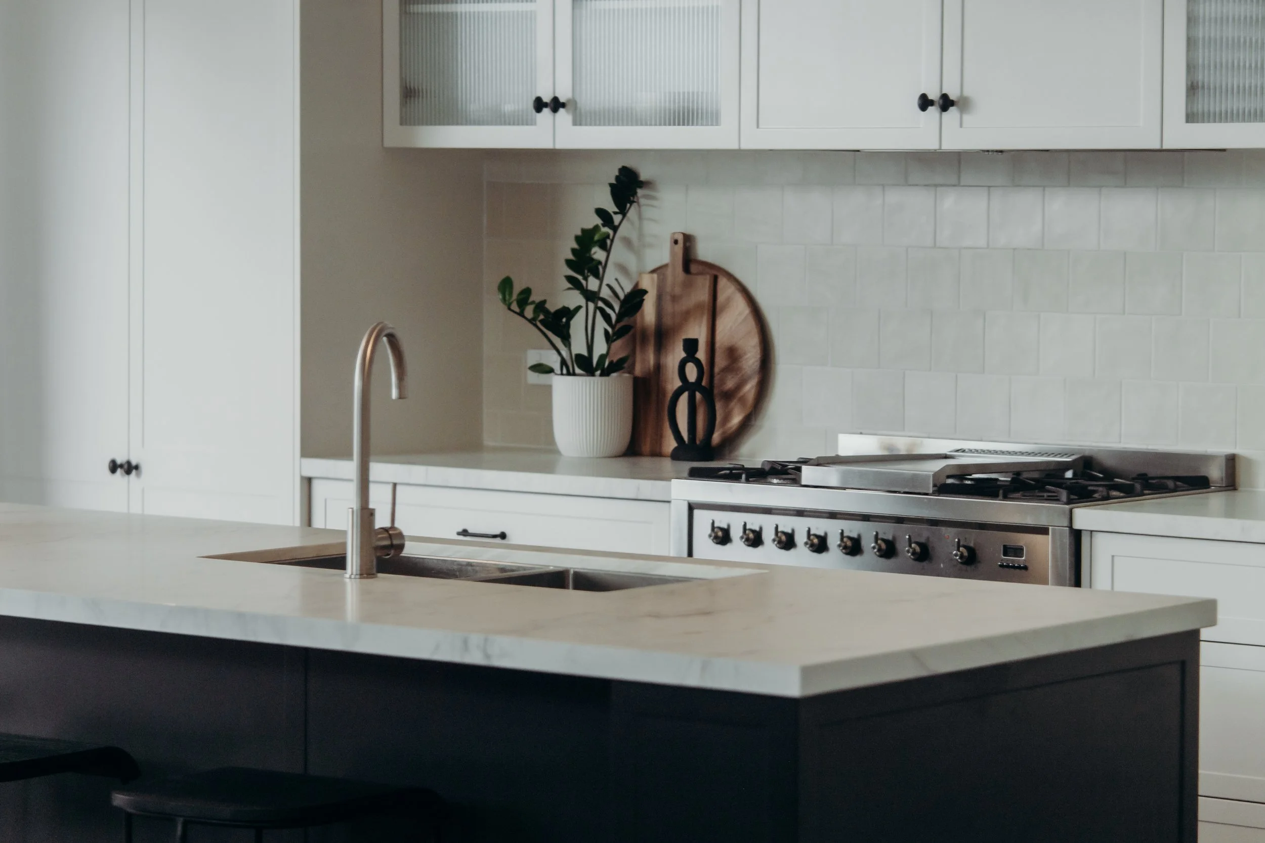 Modern kitchen with white cabinets, a black stove, a kitchen island with a marble countertop, and decorative items on the counter including a potted plant, a wooden tray, and a black sculpture.