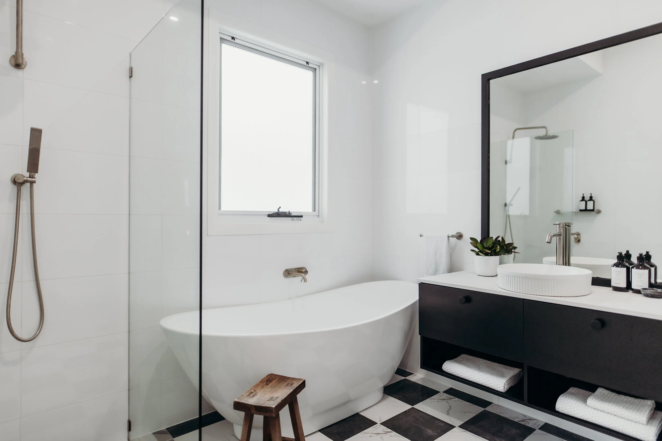 Modern white bathroom with black accents, featuring a freestanding bathtub, a window, a black vanity with a white vessel sink, a large mirror, a plant, and neatly folded towels.