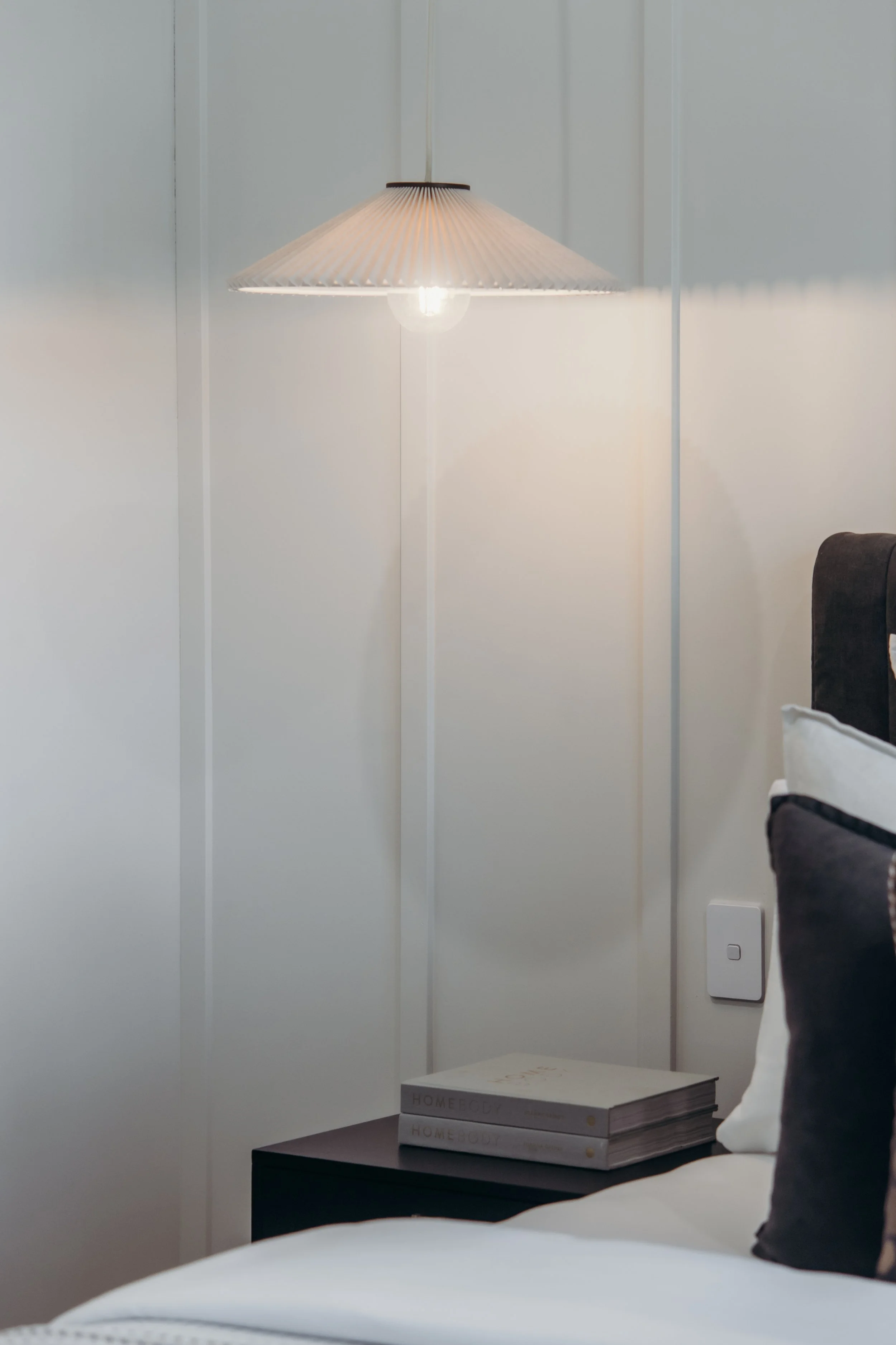 A modern bedroom corner with a white wall, a hanging pendant lamp with a paper shade, a bedside table with two stacked books titled "HOME BODY," a bed with white sheets and a black and white pillow.