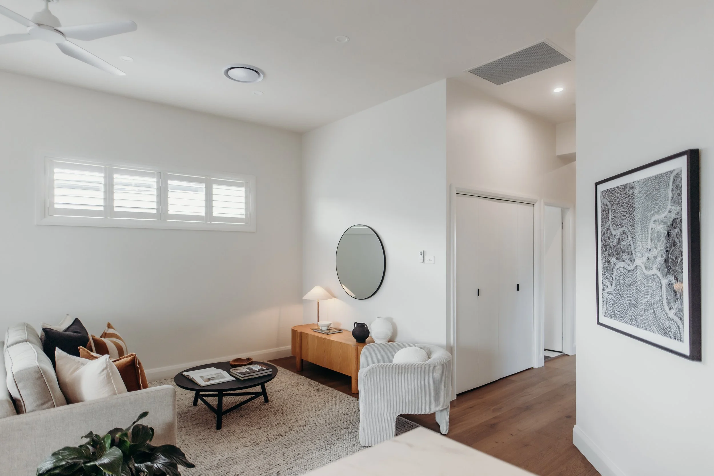 Living room with white walls, a beige sofa with pillows, a black coffee table, a white armchair, a wooden side table with a lamp and decorative vases, a mirror on the wall, a framed abstract artwork, a window with white shutters, hardwood floors, and