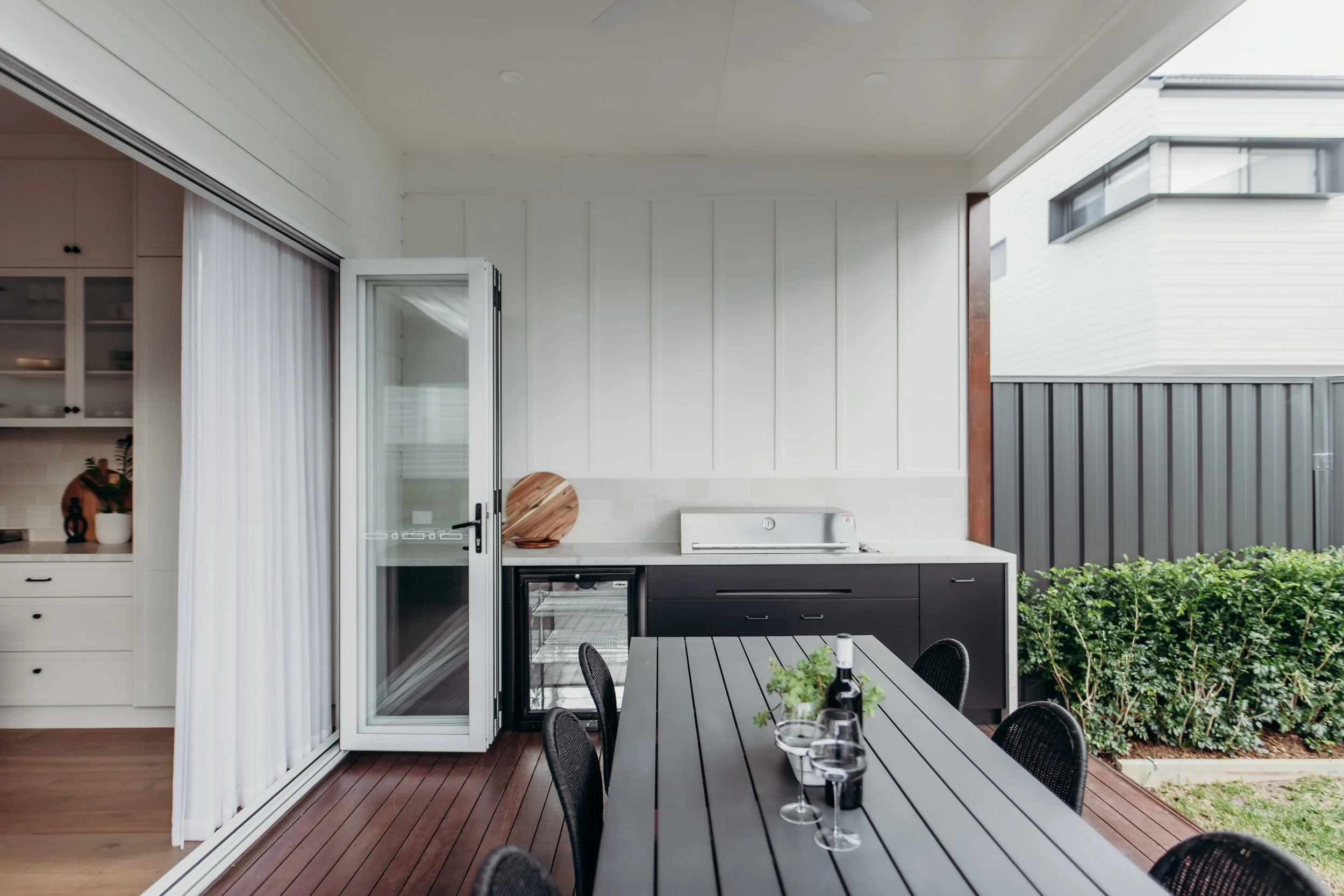 Outdoor patio area with a black dining table, six black chairs, a bottle of wine, and four wine glasses. Behind the table is a built-in outdoor kitchen with a small refrigerator, a sink, and black cabinetry. There are green bushes along a gray fence,
