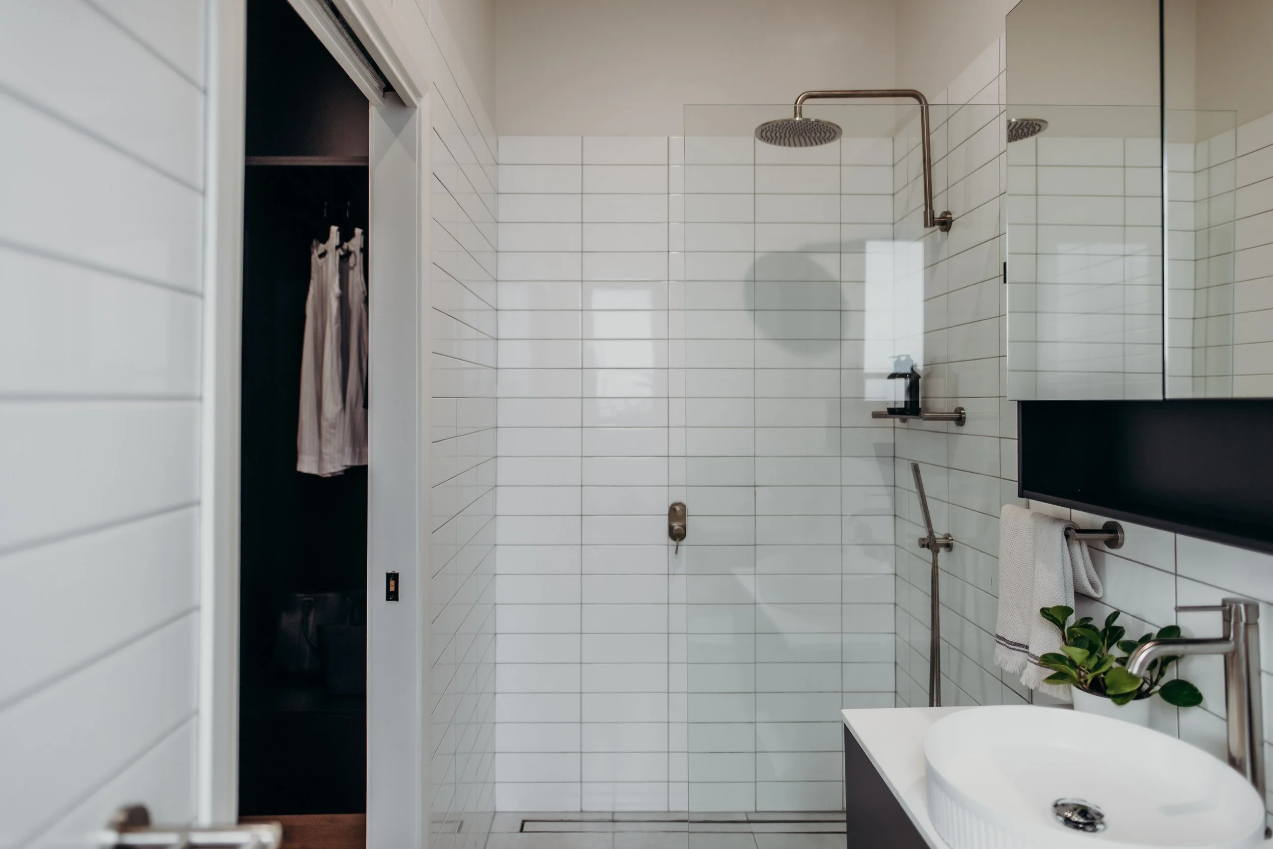 Modern white tiled bathroom with glass shower enclosure, wall-mounted showerhead, white sink, and mirror cabinet, with a towel bar and green plant.