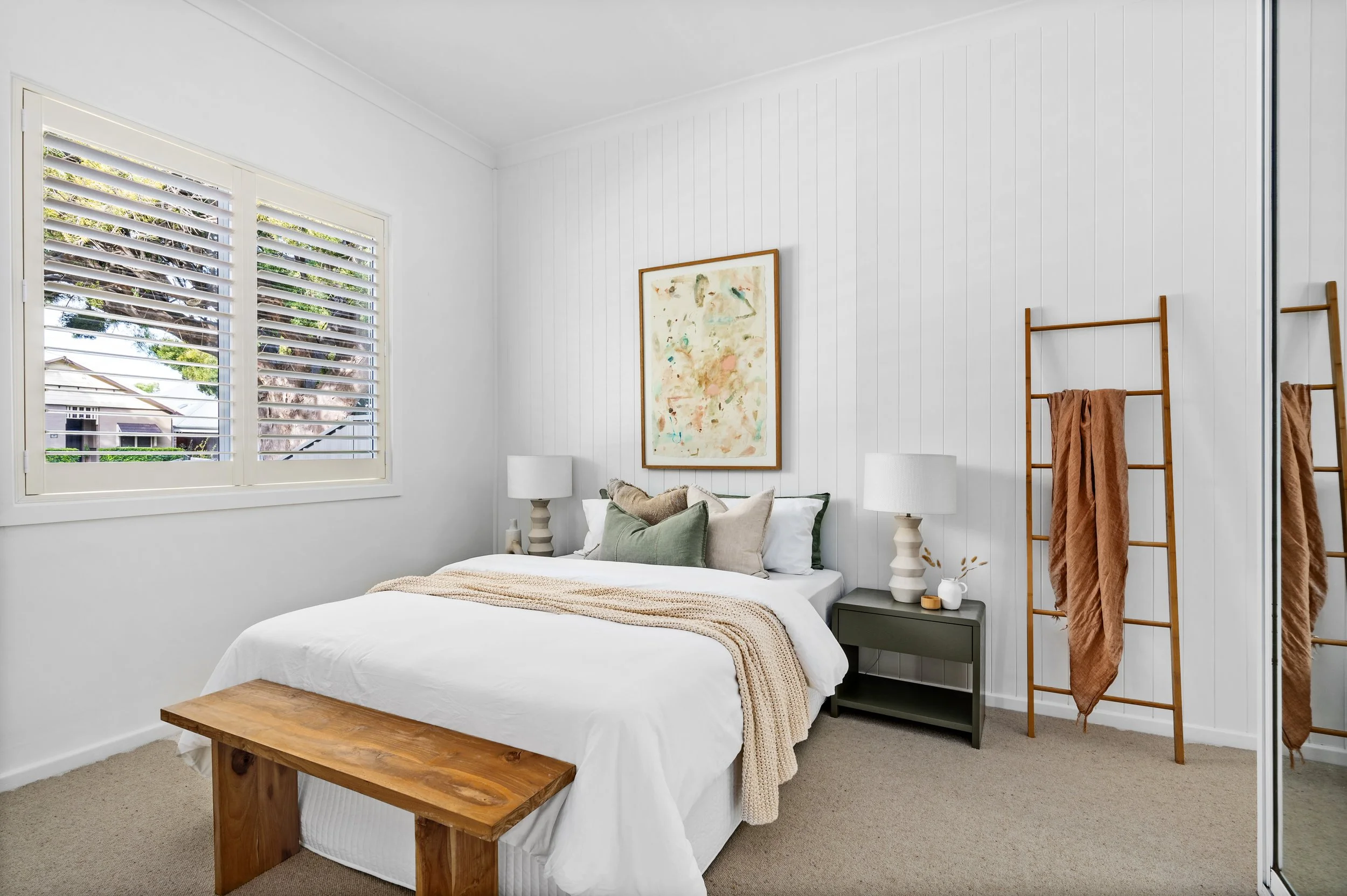 Bright bedroom with white walls, a large window with white shutters, a bed with white bedding and decorative pillows, two matching white lamps on dark green nightstands, a wooden bench at foot of bed, wall art above bed, and a wooden ladder with brow