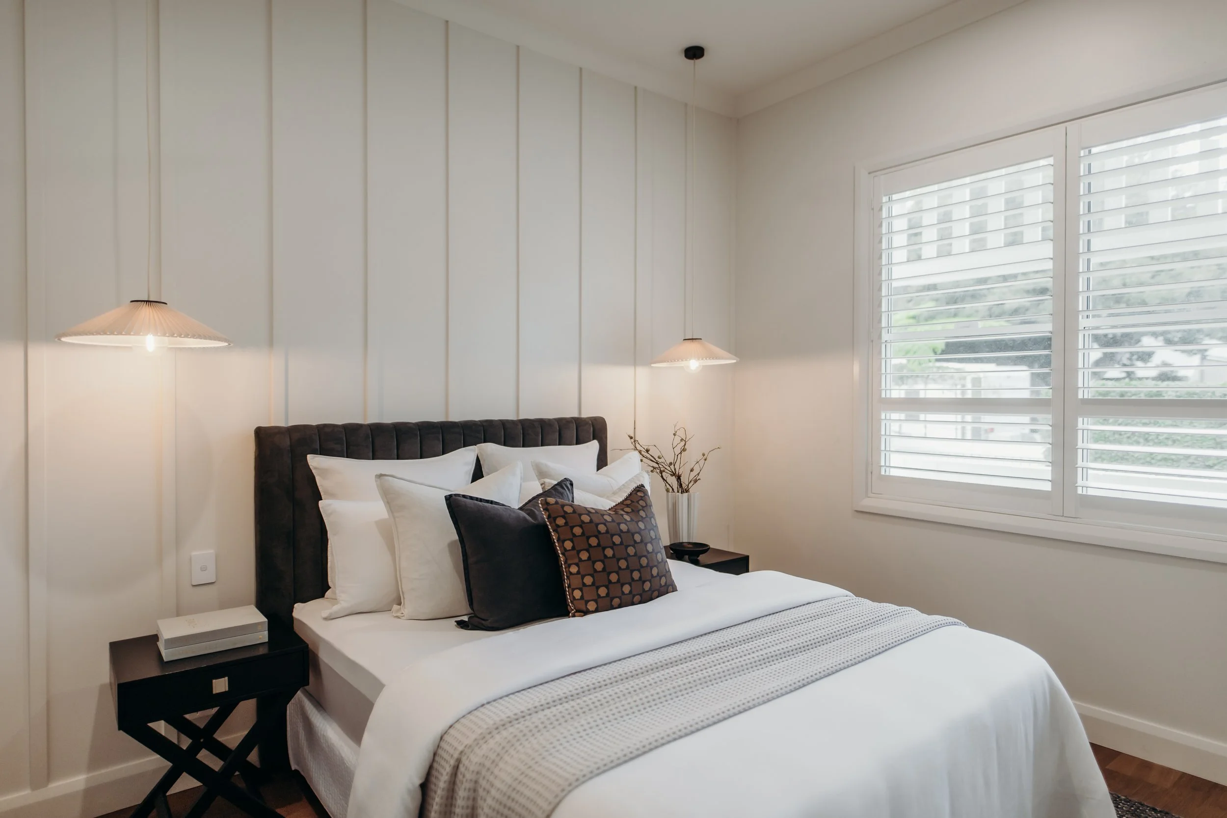 A neatly made bed with white sheets and pillows, dark headboard, side tables with lamps, and a window with white blinds in a minimalist bedroom.