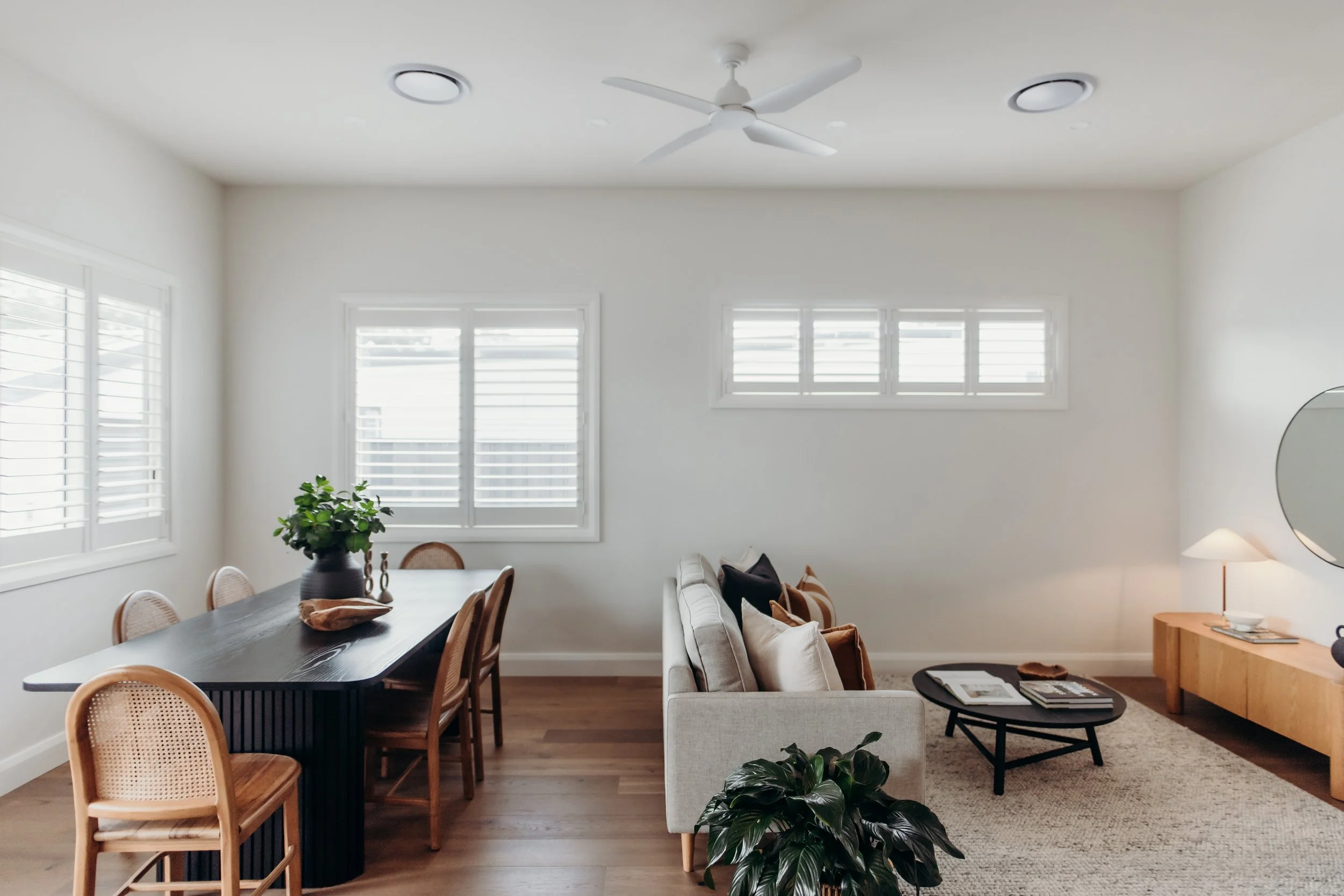 Bright living room and dining area with white walls, wooden flooring, sofa with cushions, coffee table, wooden TV stand, round mirror, side lamp, and large windows with shutters.
