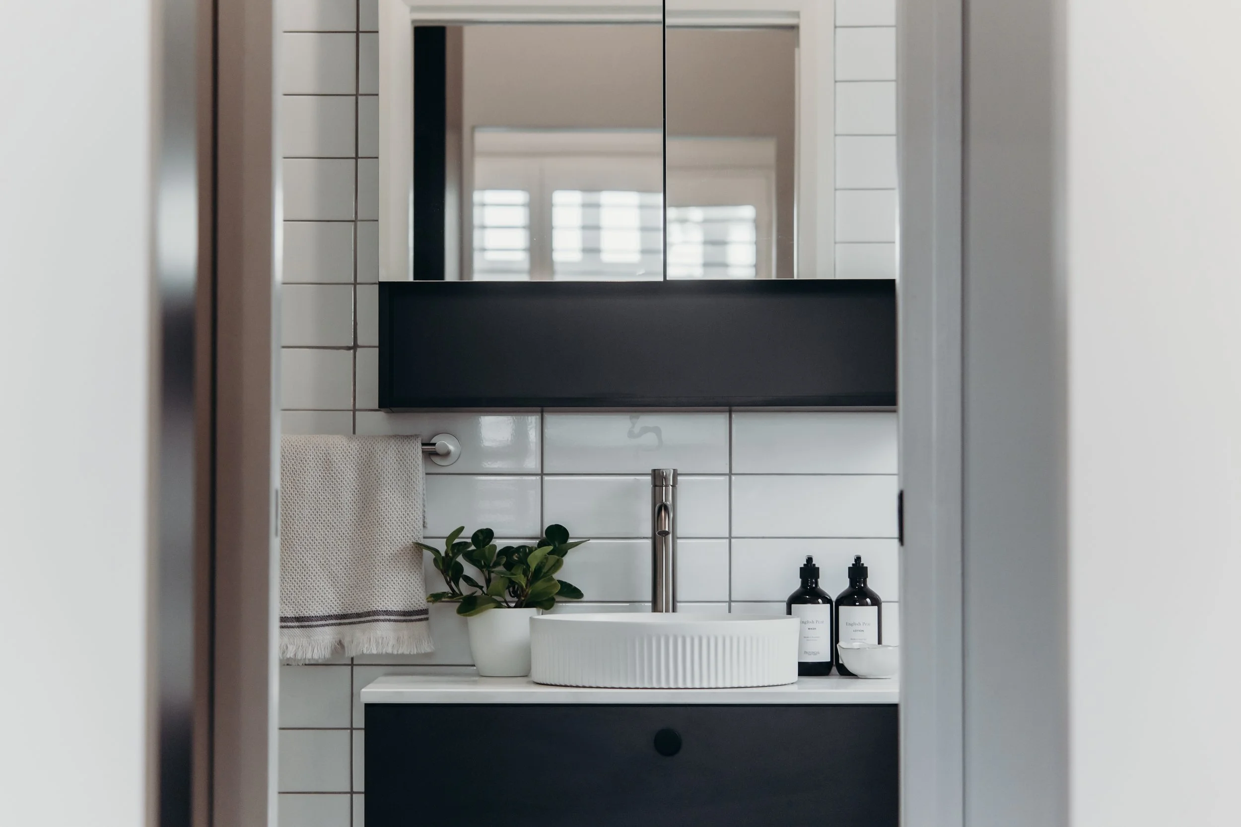 Modern bathroom with white tiled wall, black vanity, round white vessel sink, metallic faucet, mirror cabinet, potted plant, two black soap bottles, hand towel, and window reflections