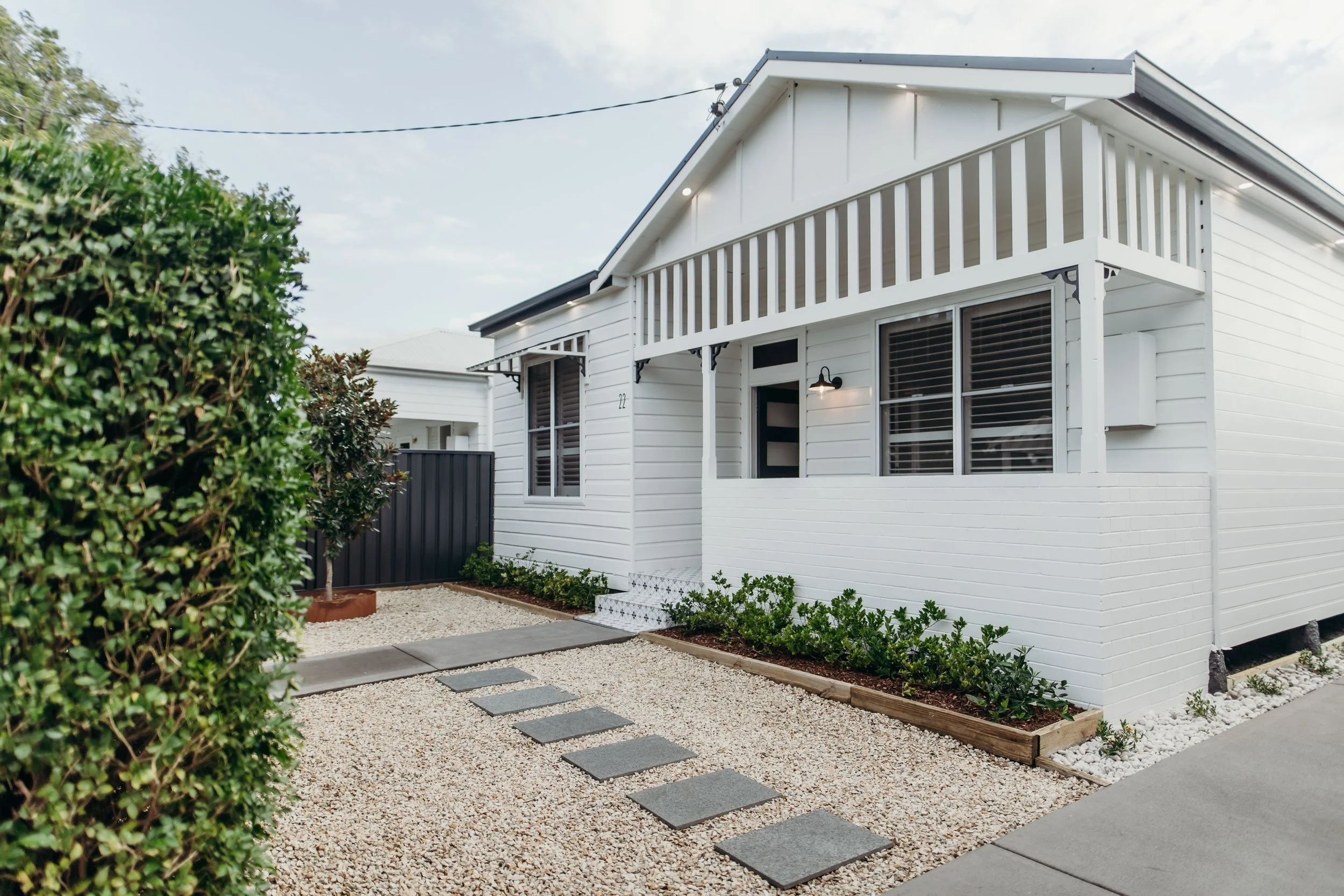 A modern white house with a small front porch, black trim, and a balcony. The yard has a gravel pathway with stone slabs and small landscaping plants.
