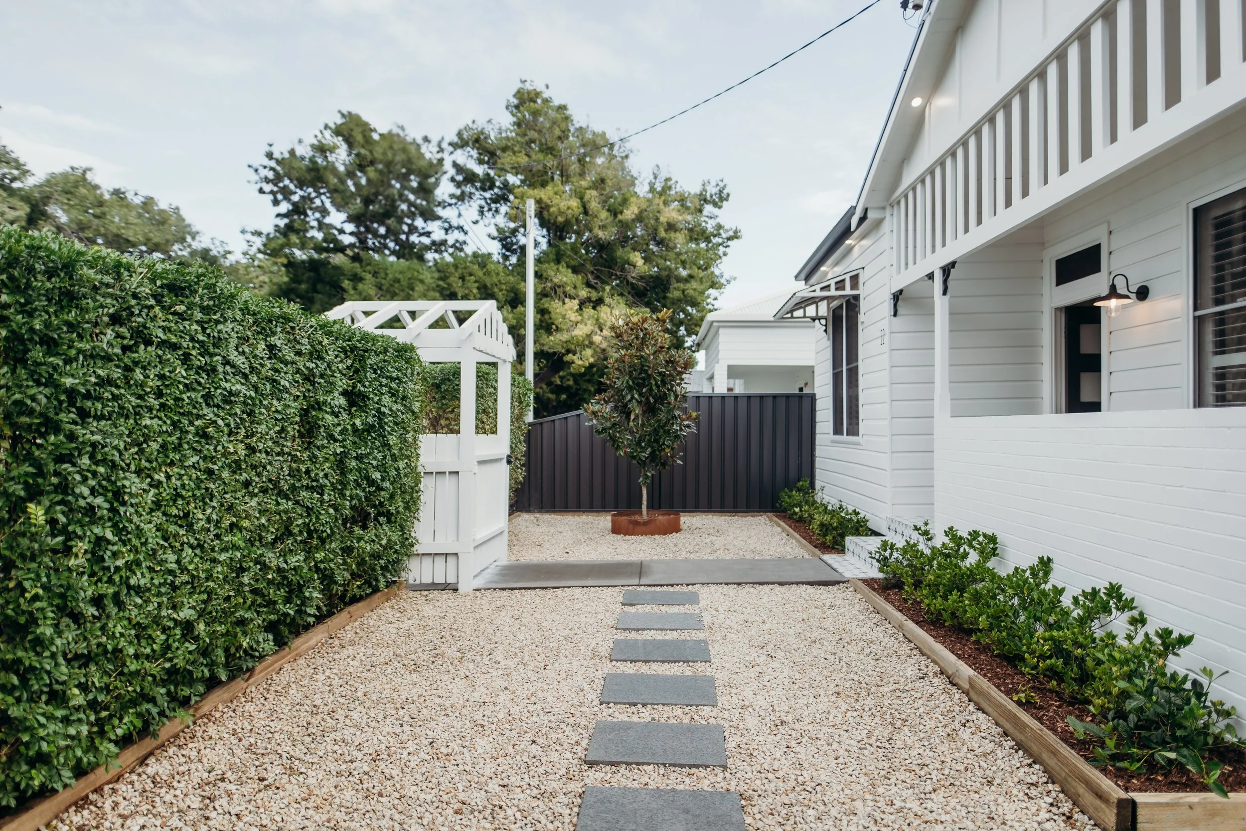 A modern backyard with a gravel pathway, small rectangular stepping stones, green bushes on the right side, a white picket gate on the left side, and a white house with black trim and a porch light on the right. In the background, a small tree and a
