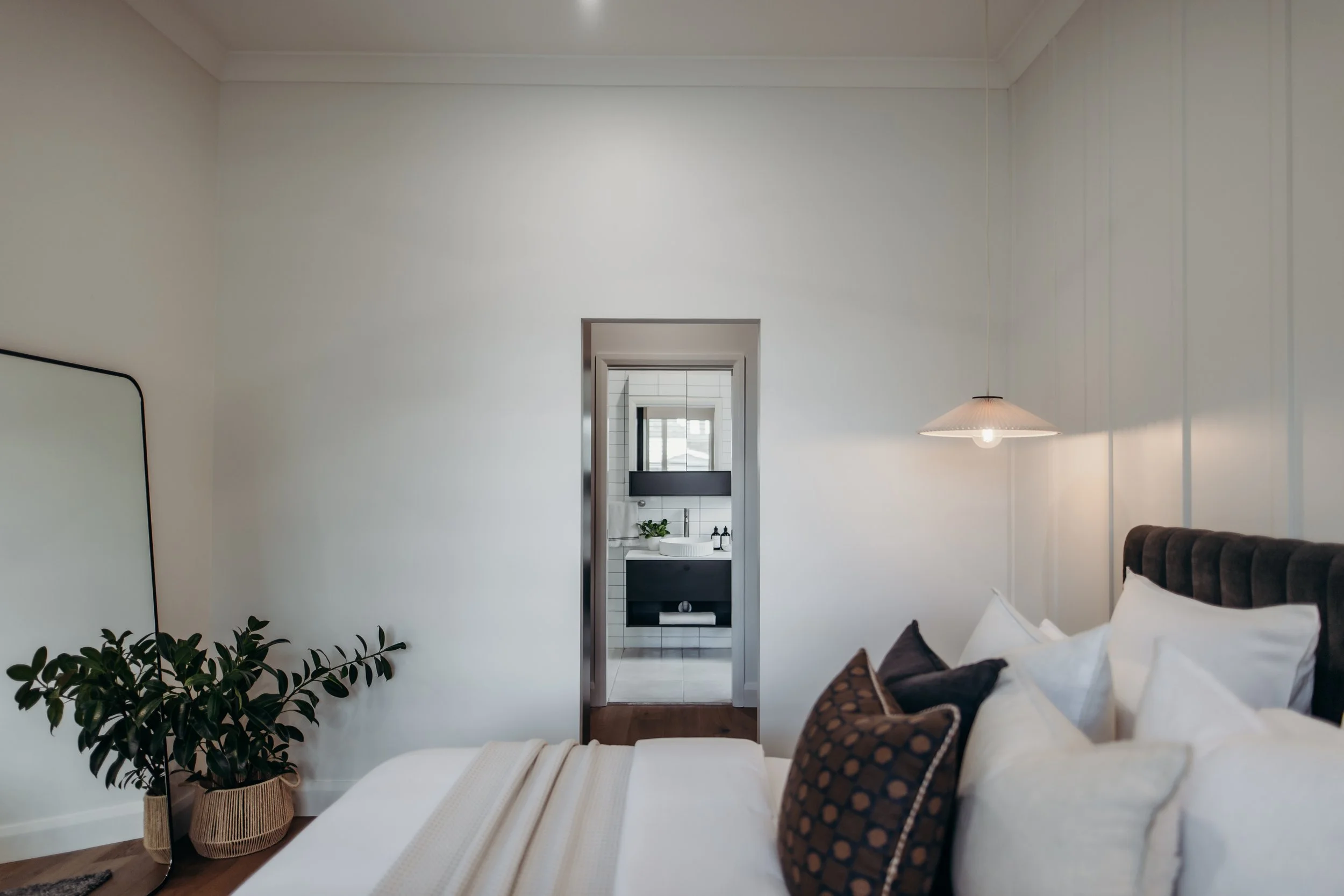 A bedroom with a bed, pillows, a wall-mounted light, and a view into a bathroom through a doorway.