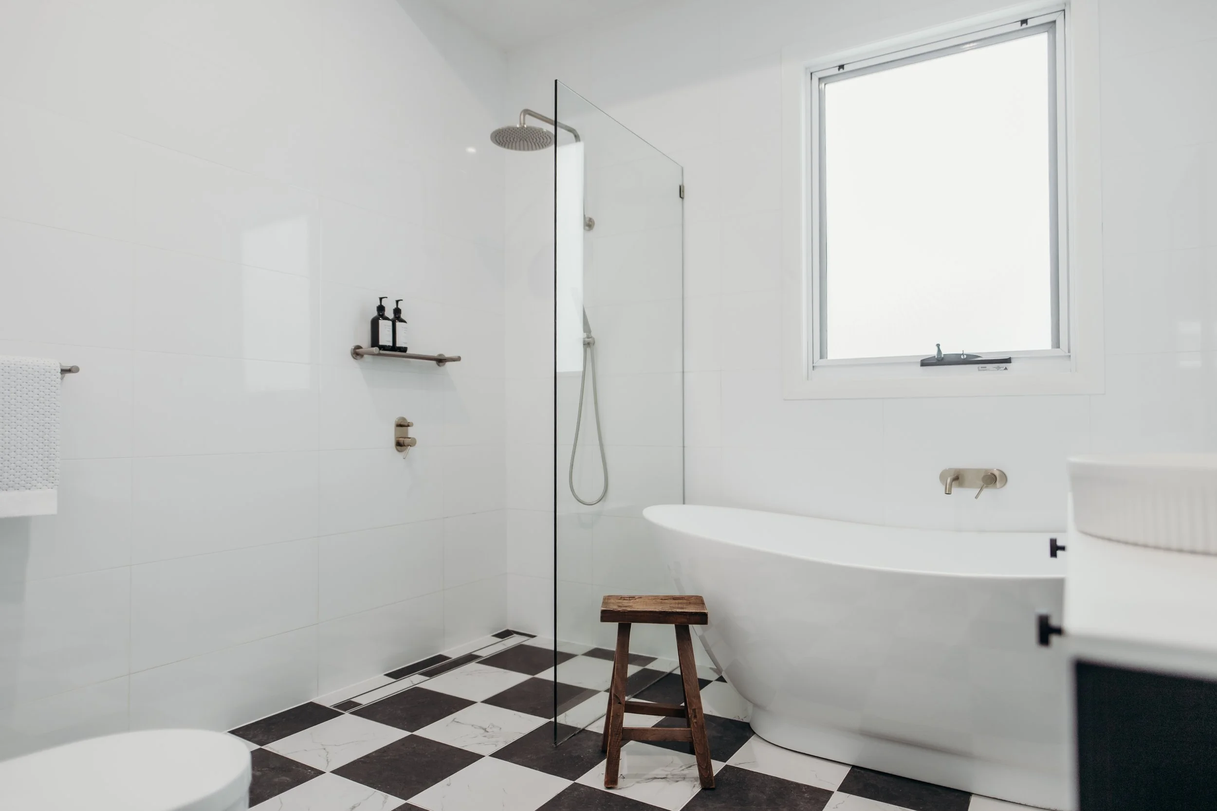 Minimalist bathroom with a walk-in shower, a white bathtub, black and white checkered floor tiles, a window, and a small wooden stool.