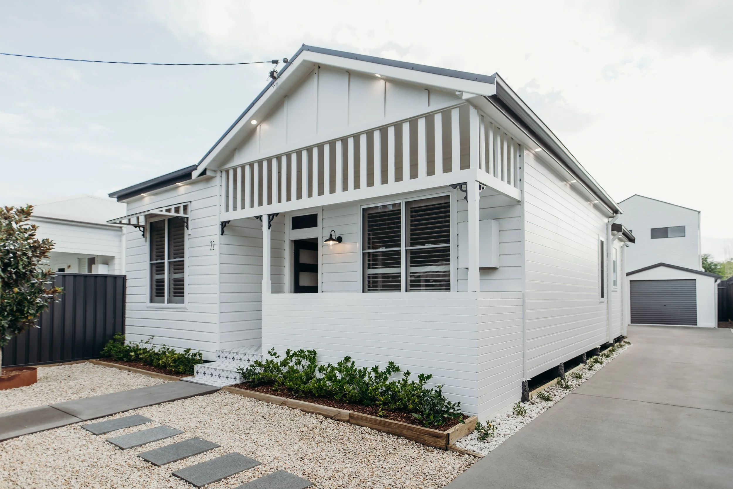 Modern white house with a small front porch and second-floor balcony, surrounded by a minimalist garden with white gravel and green shrubs.