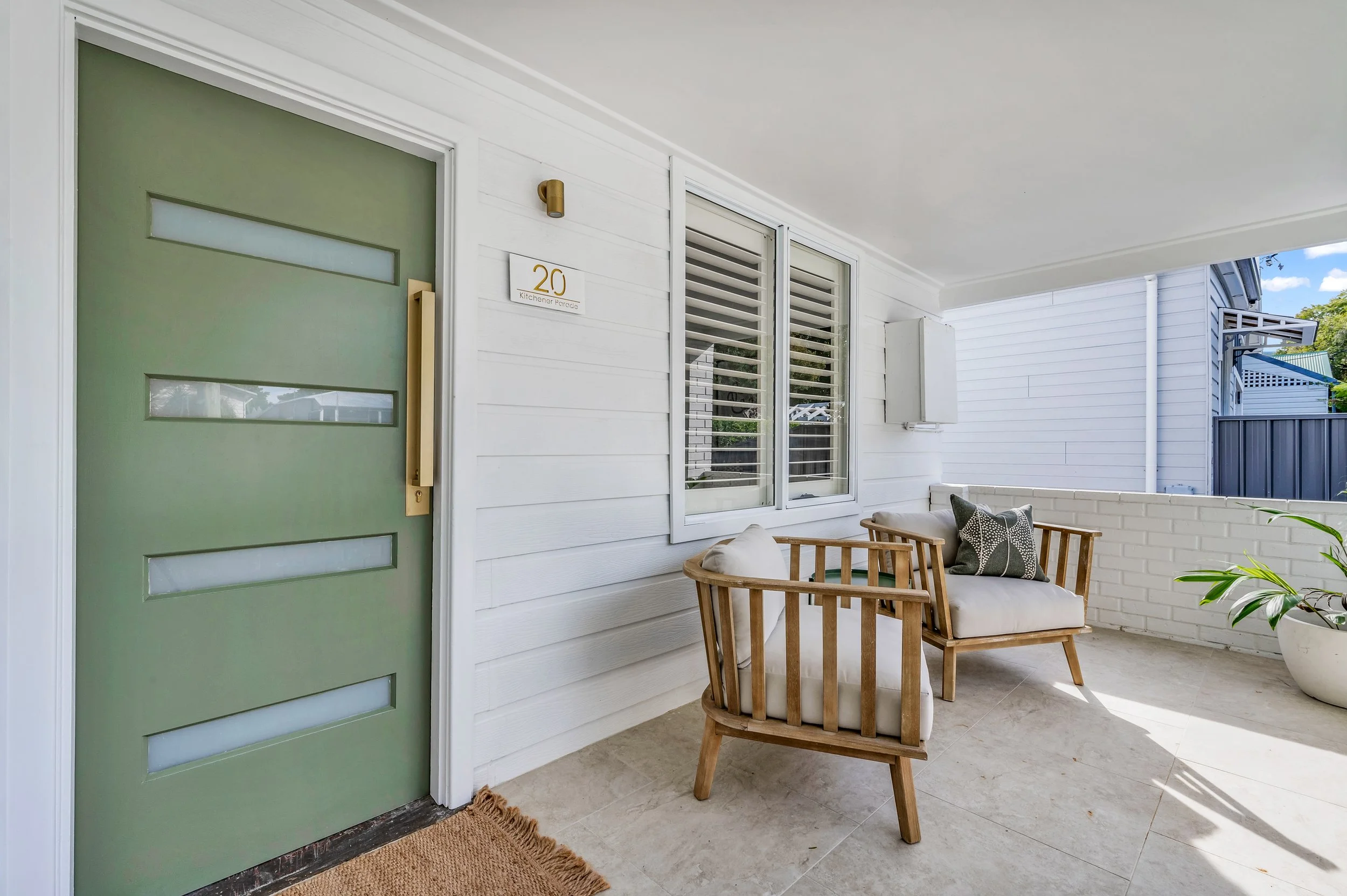 Front porch with green door, white house siding, two windows with shutters, seating area with wooden chairs and cushions, potted plant, house number 20, and a small wall-mounted electrical box.