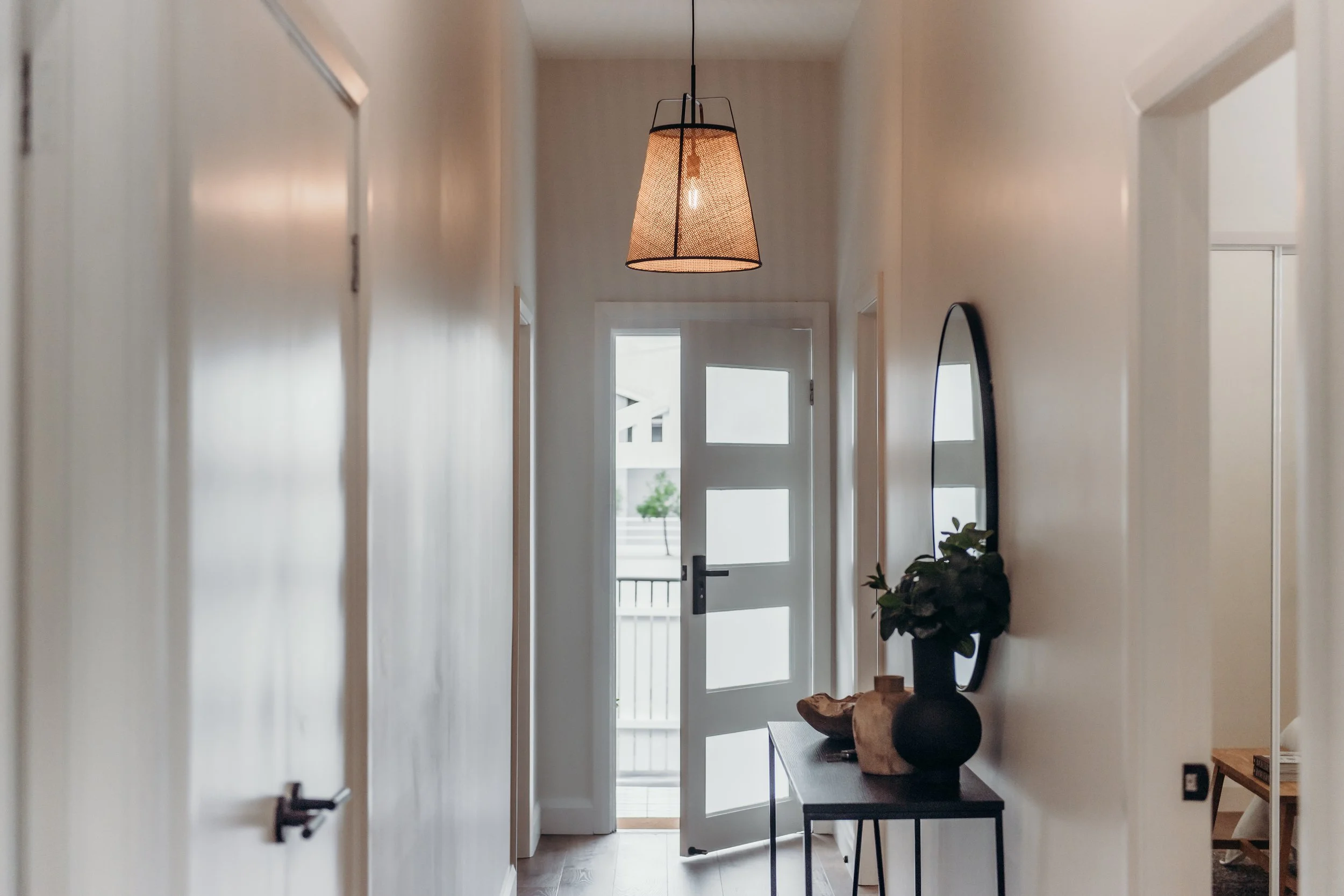 A modern, minimalist hallway with a light fixture hanging from the ceiling, a door with glass panels open to a balcony, and a console table with decorative vases and a mirror on the wall.
