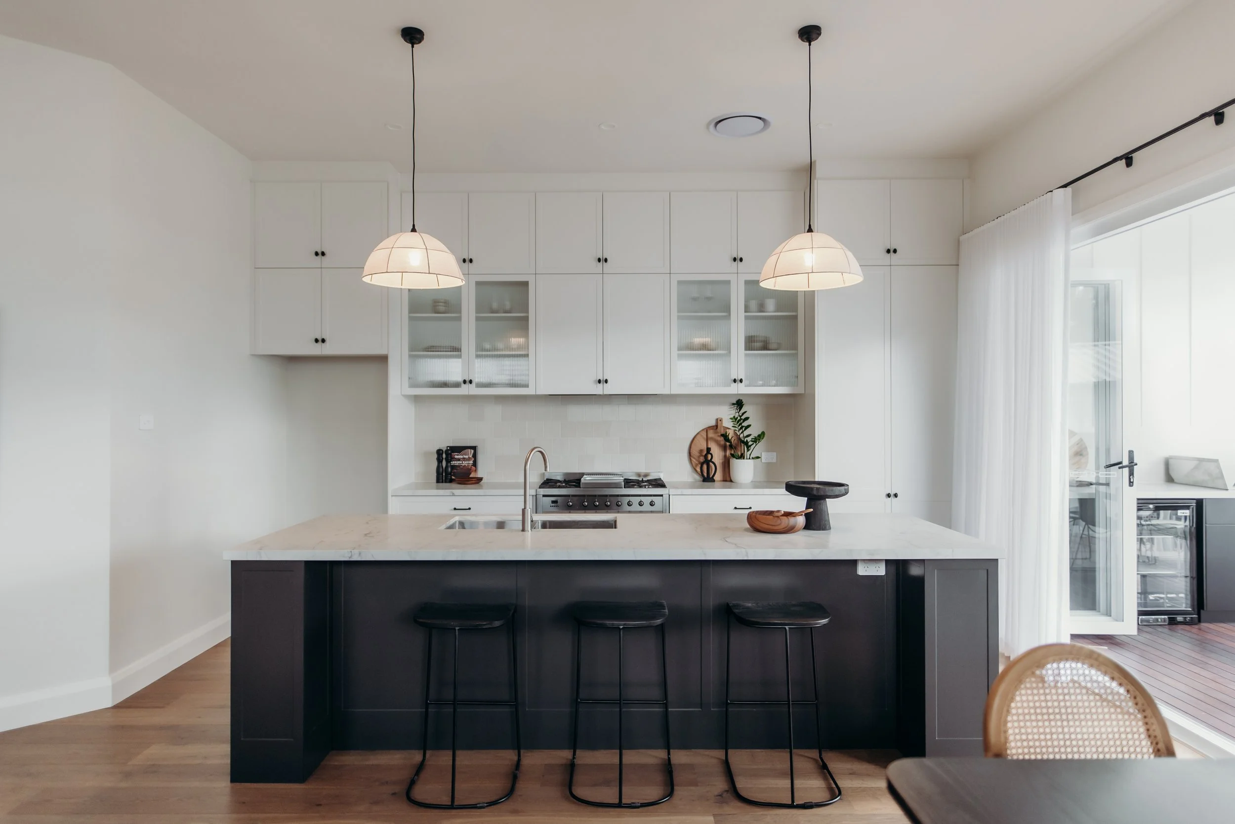 Modern kitchen with white cabinets, black island, and pendant lights, featuring a marble countertop, bar stools, and a sliding glass door with white curtains.