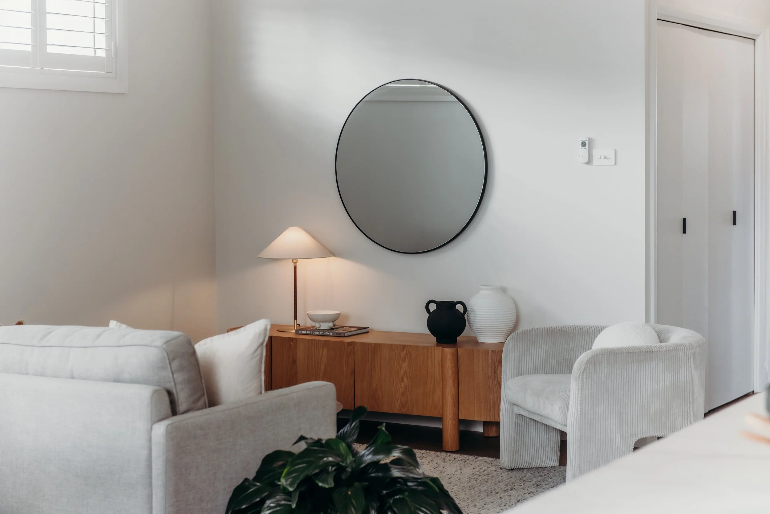 Modern living room with white furniture, a wooden console table, a round mirror, a table lamp, and decorative vases.
