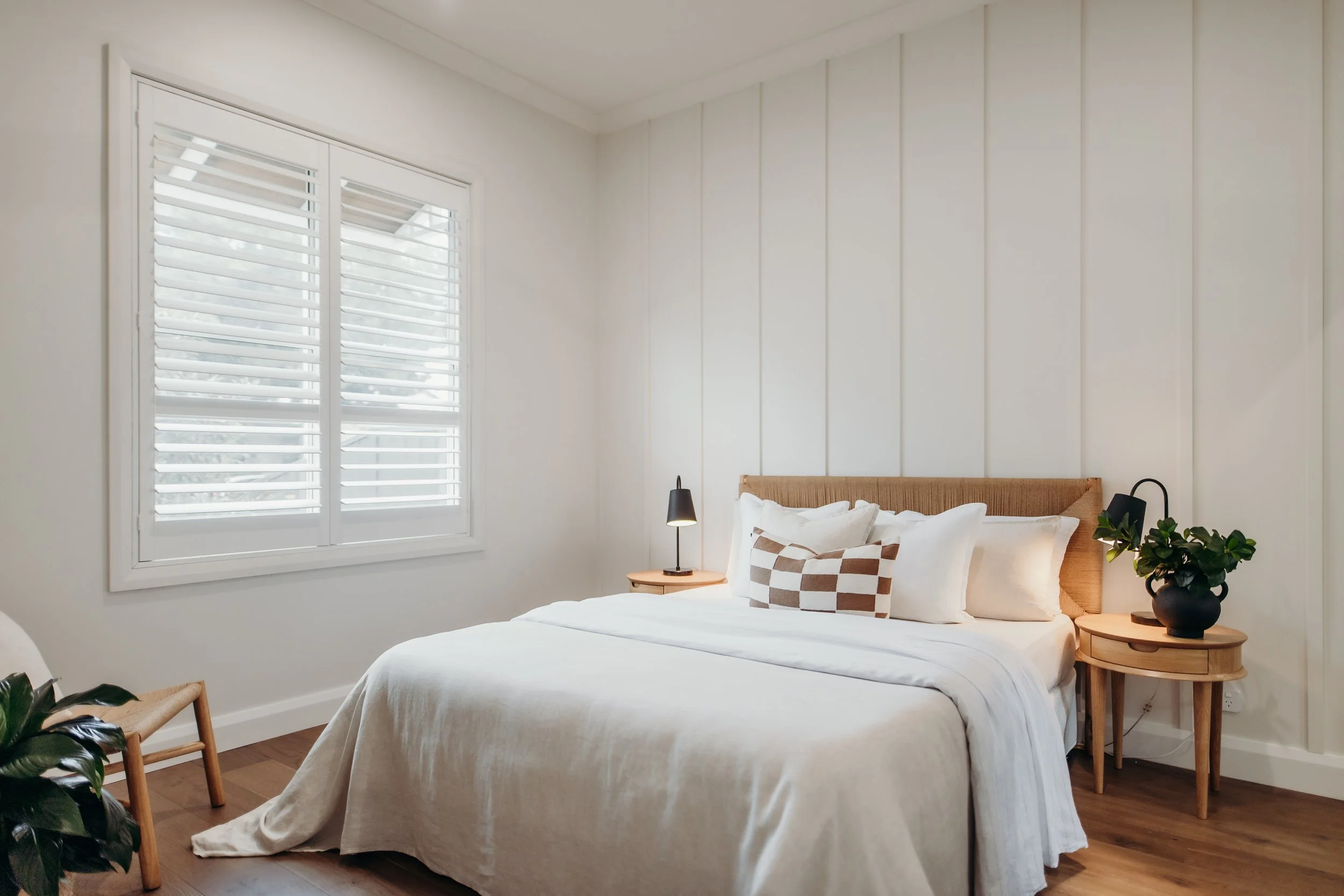 A cozy bedroom with a white bed, beige headboard, pillows, black bedside lamps, a small wooden table, a black vase with greenery, and a window with white shutters.