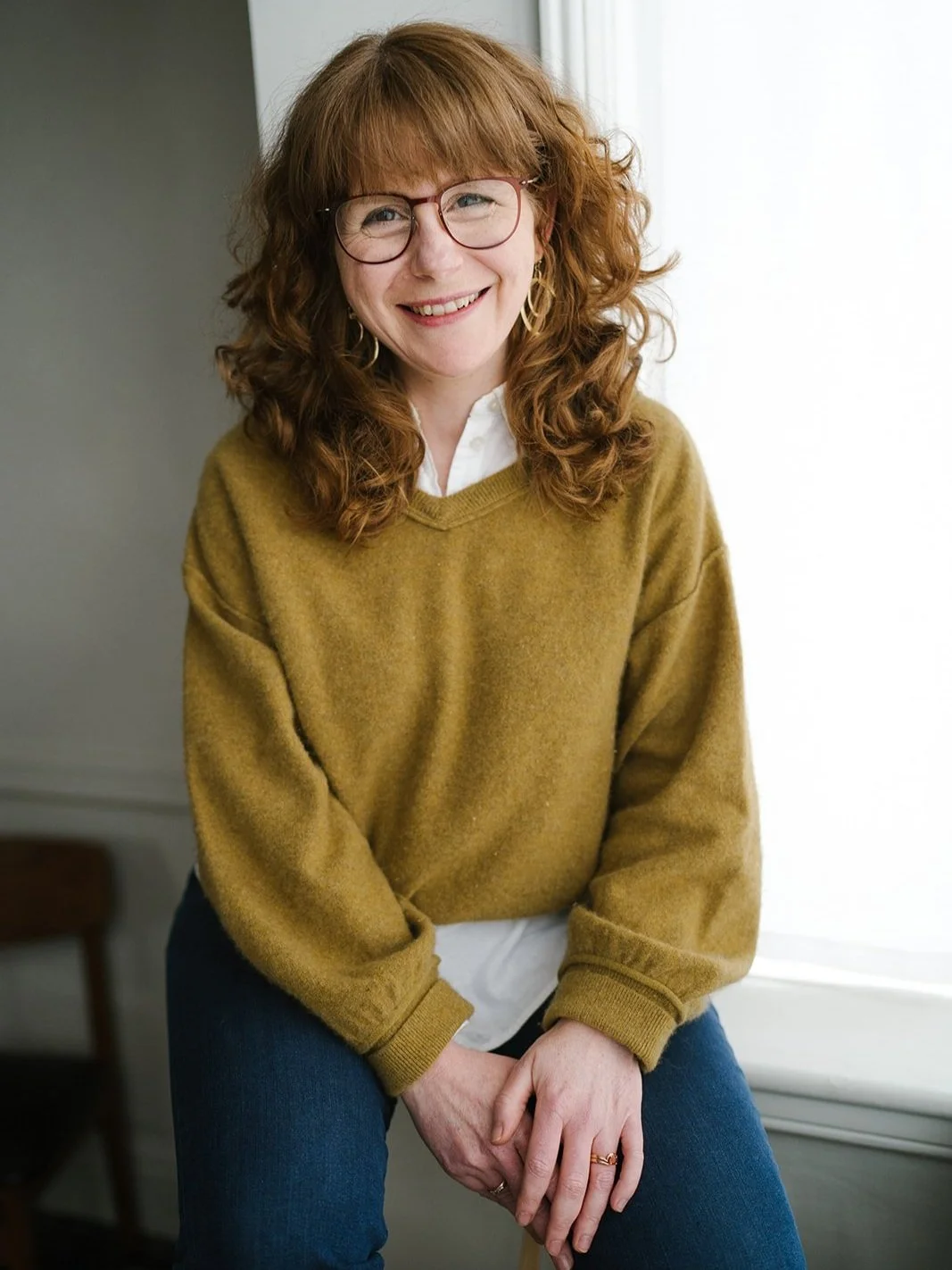 Cassandra Jackson-Baker sitting near a window and smiling. She has red curly hair and glasses.