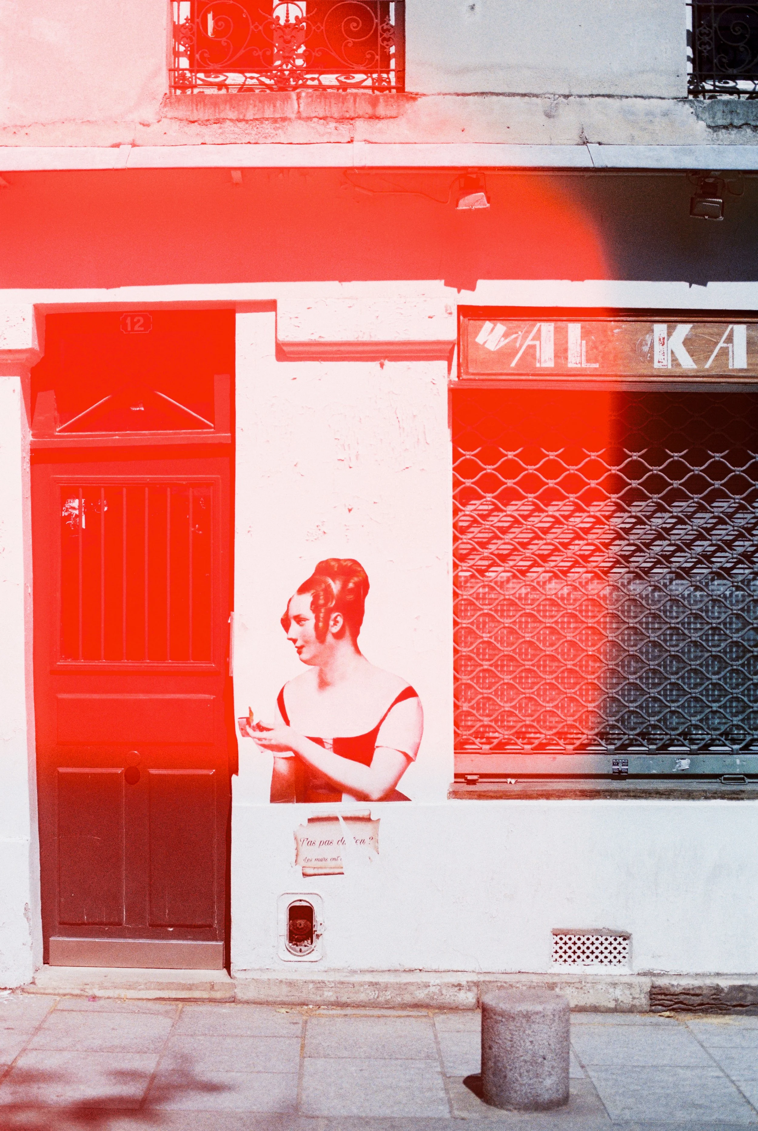 A black and white mural of a woman with vintage-style hair and dress, looking at a phone, on a white wall next to a window with a metal grill. The scene is overlaid with a red filter.