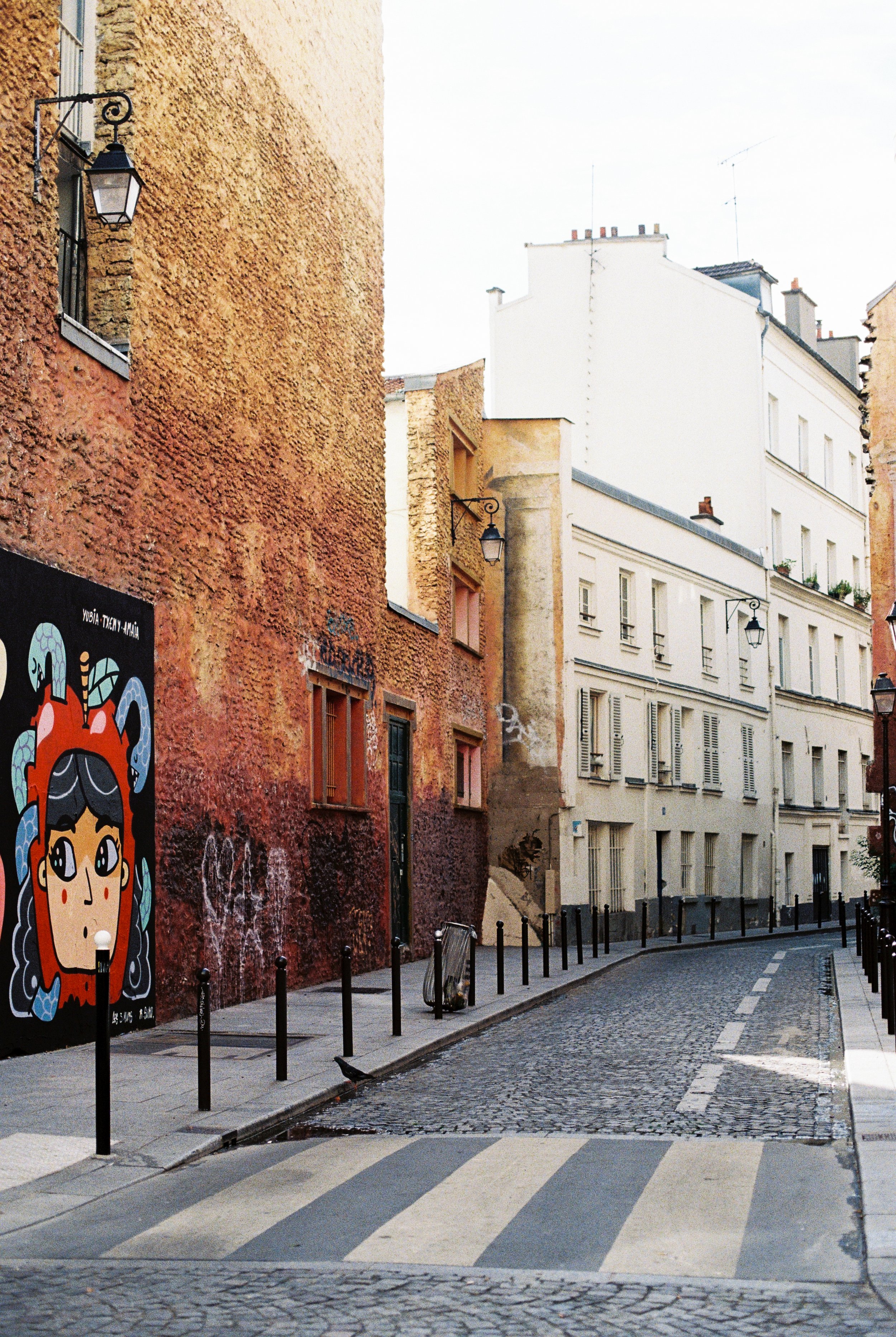 A cobblestone street in a European city, lined with colorful buildings, street lamps, a bicycle, and graffiti art on one wall.