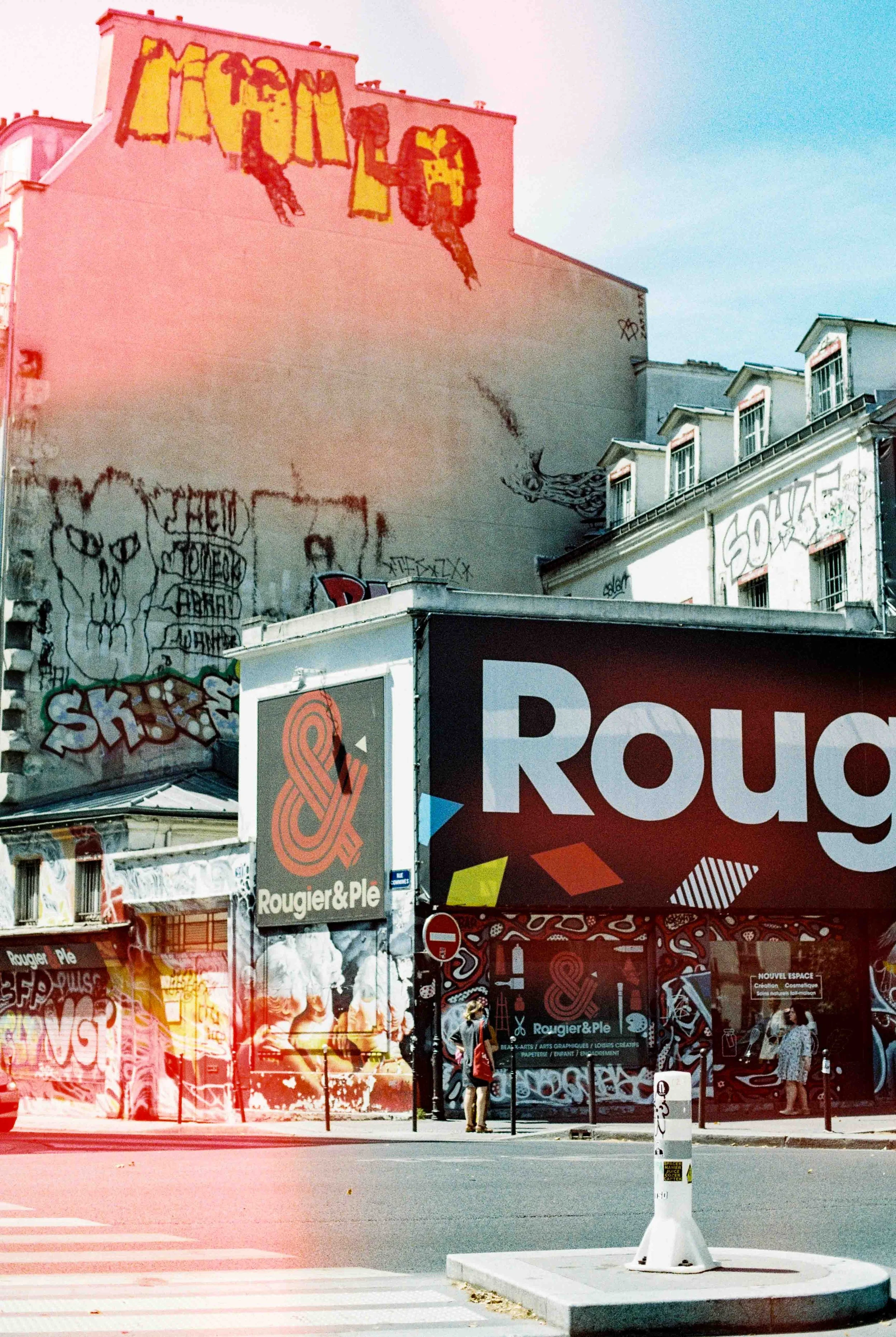 Street scene with graffiti-covered walls, a large black and red sign for Rougier & Ple, and people walking on the sidewalk. The buildings are decorated with various graffiti and street art.