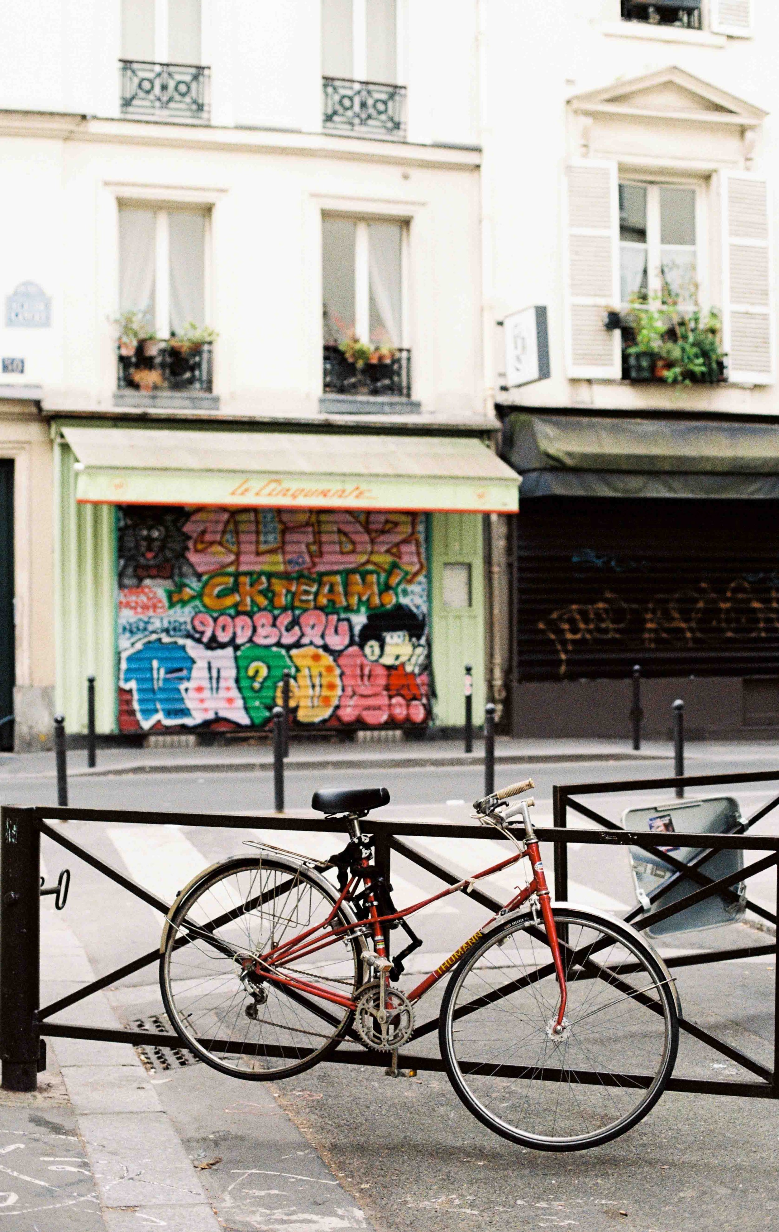 People using shared bikes on a city street near a cafe or shop with outdoor seating and parked luggage.