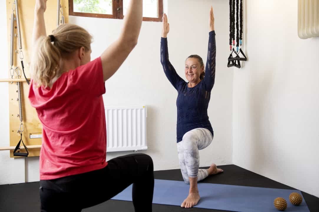 En kvinna utför yogaövning på en matta medan en annan kvinna hjälper och guidar henne i ett träningsrum.