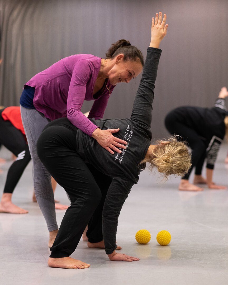 En kvinna hjälper en person att utföra en yogaövning i en gruppsportstudio.