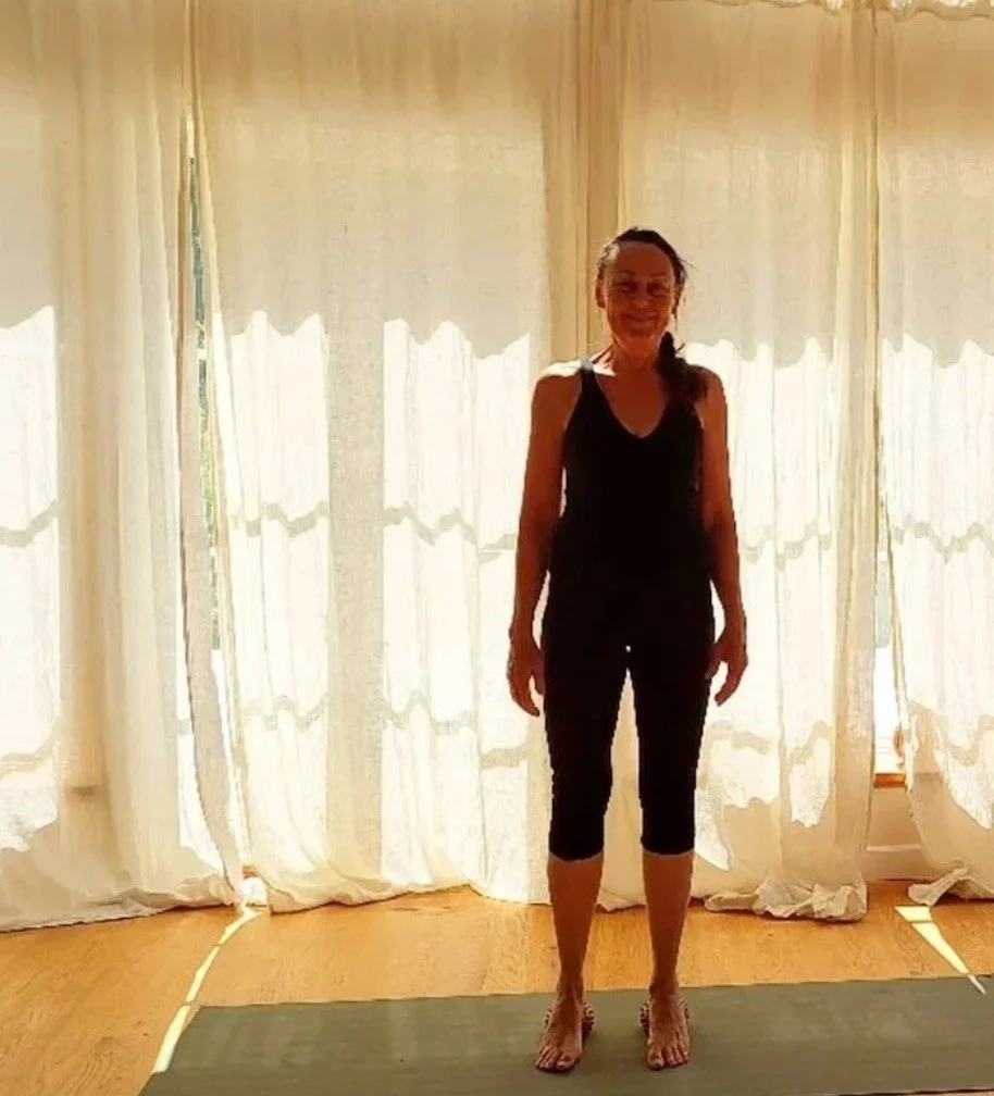 En kvinna står på en matta i ett rum med ljusa gardiner bakom sig och ler.