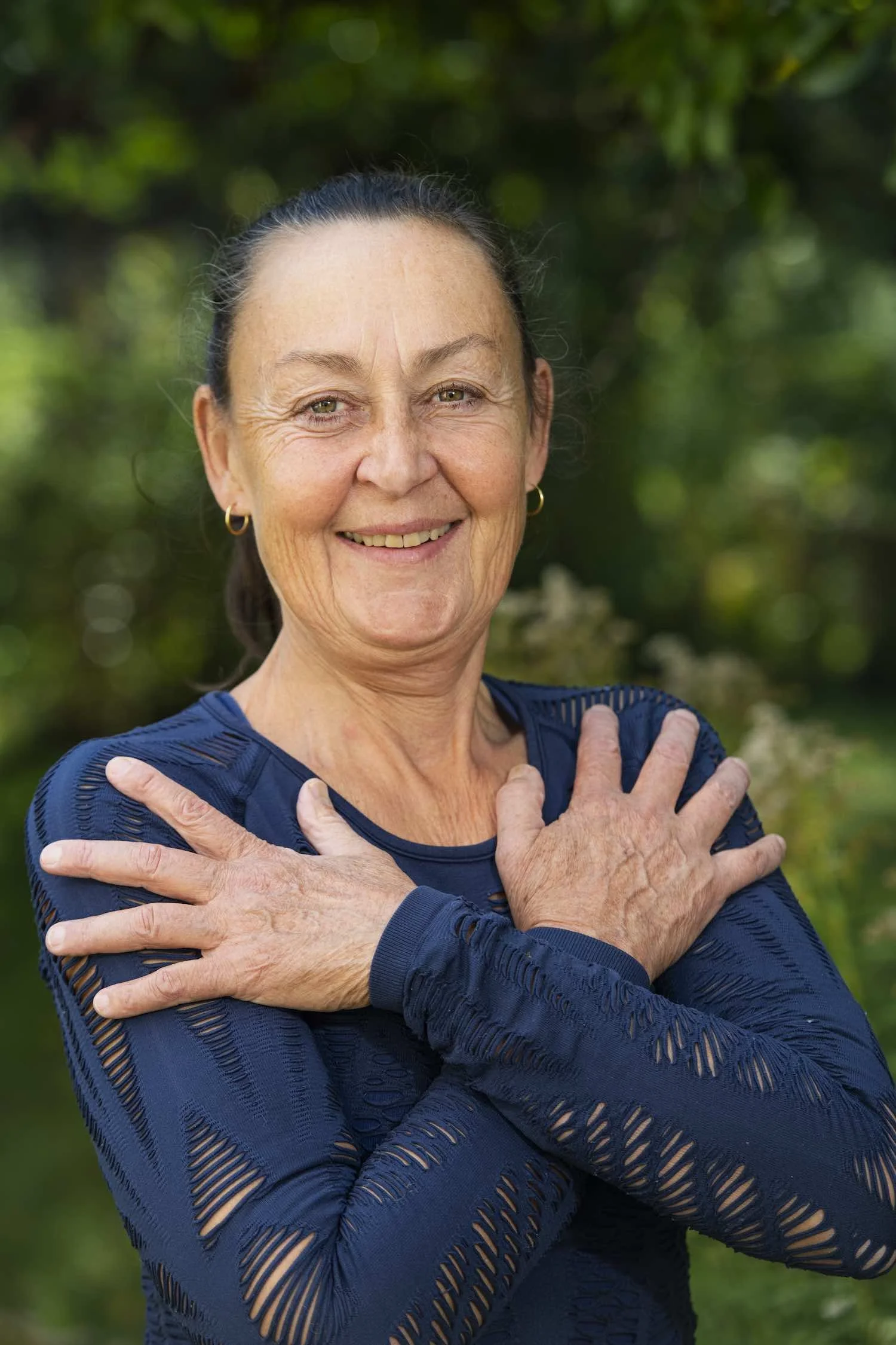 Cecilia Douglas bär en långärmad mörkblå tröja med utskurna mönster, står utomhus med grönt bladverk i bakgrunden.