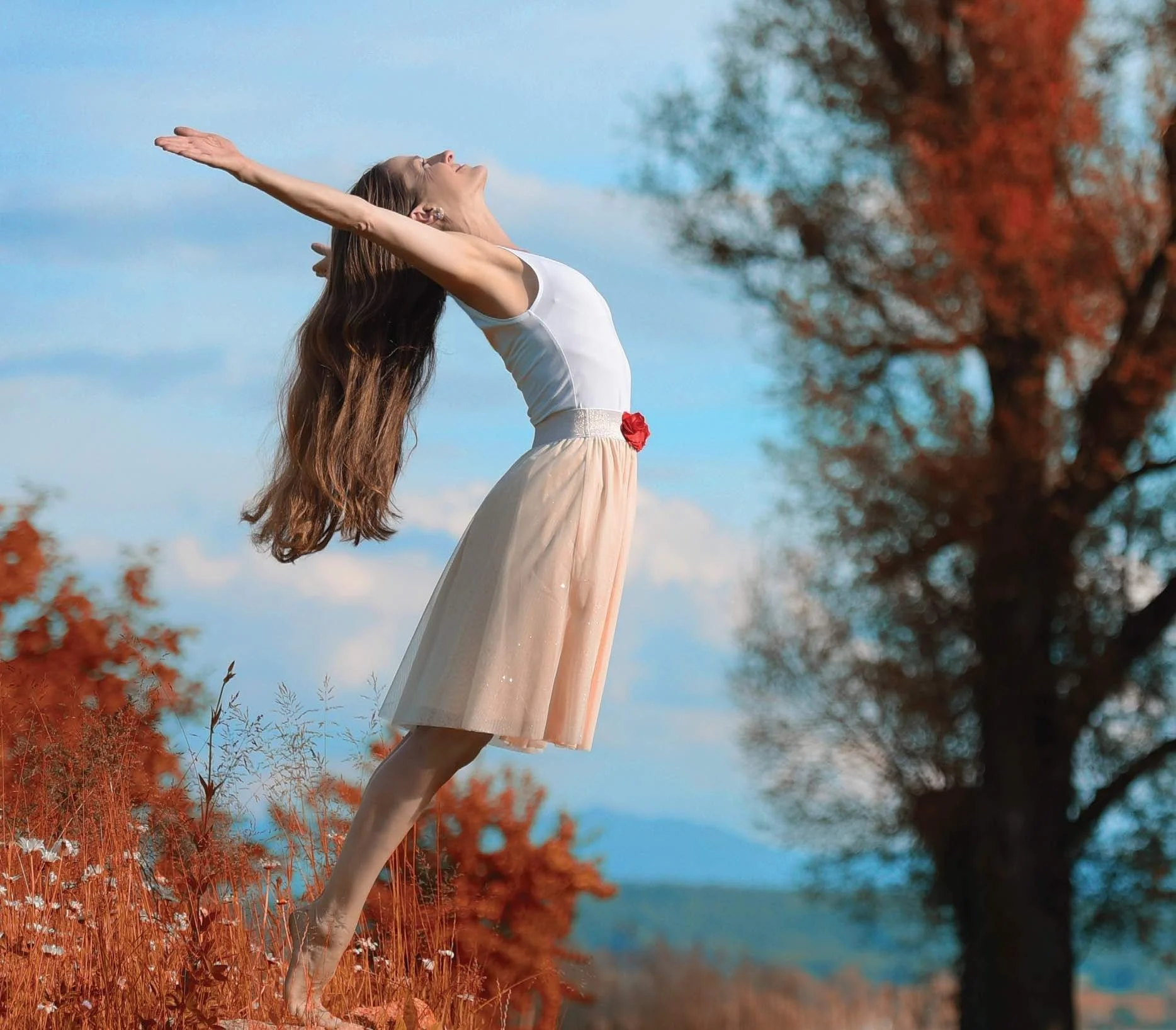 En kvinna står på ett fält med orange-lätter träd i bakgrunden, med armarna utsträckta och huvudet vänt mot himlen.