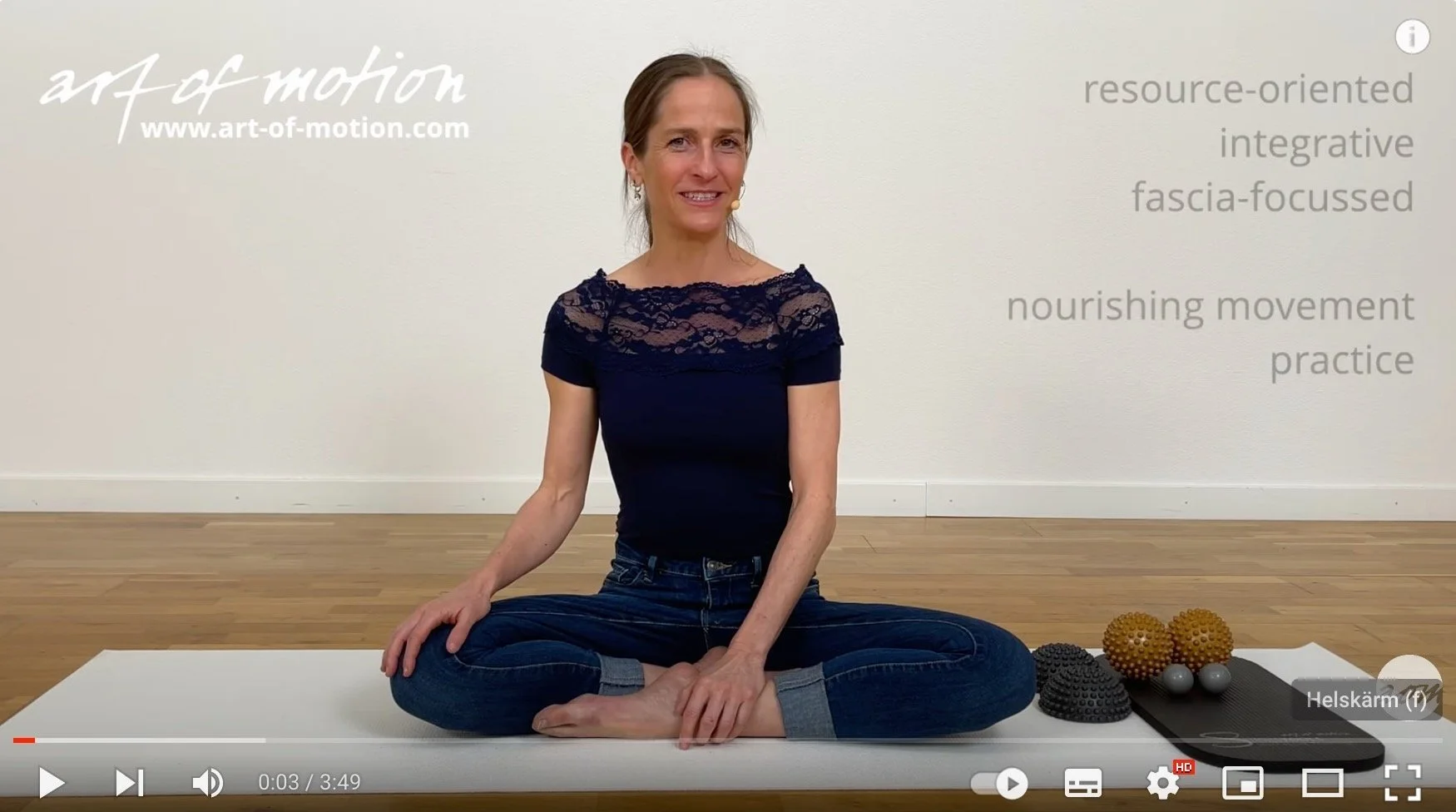 Karin Gurtner sitter på golvet i korslagda ben framför en vit vägg. Runt henne finns träningsredskap som massagebollar och en matta.